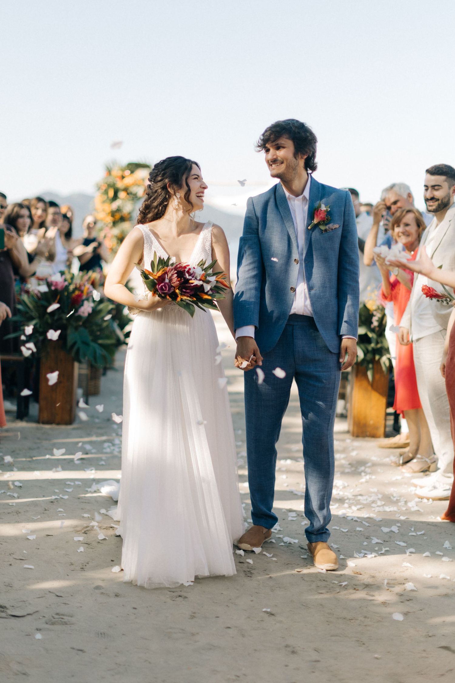 Newlyweds walking down beach aisle while guests throw petals in celebration.