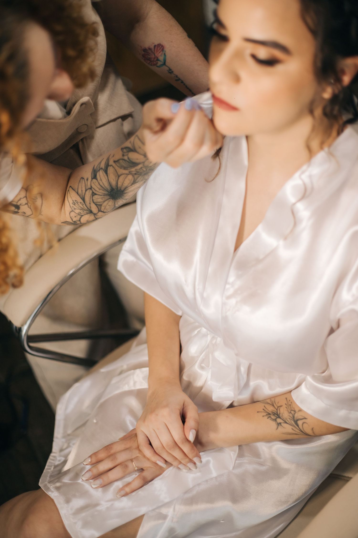 Makeup artist applies cosmetics to client wearing white silk robe with visible floral tattoos.