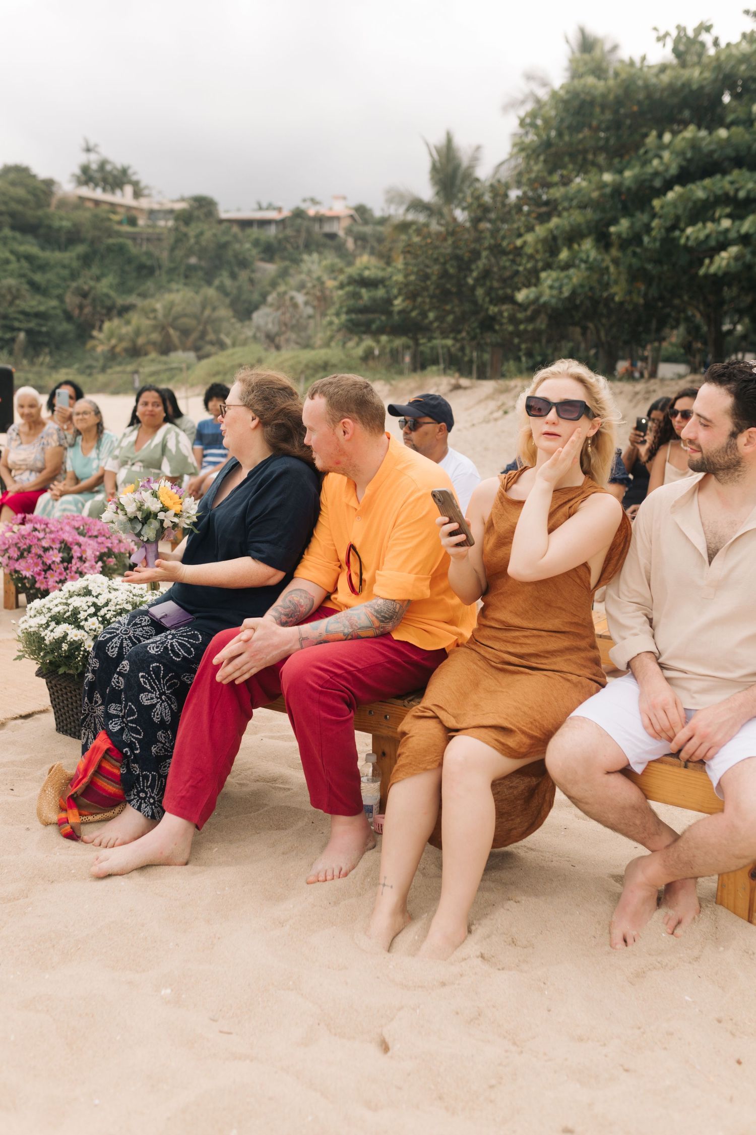 Wedding guests sitting on a beach with colorful attire and floral decorations on a sunny day.