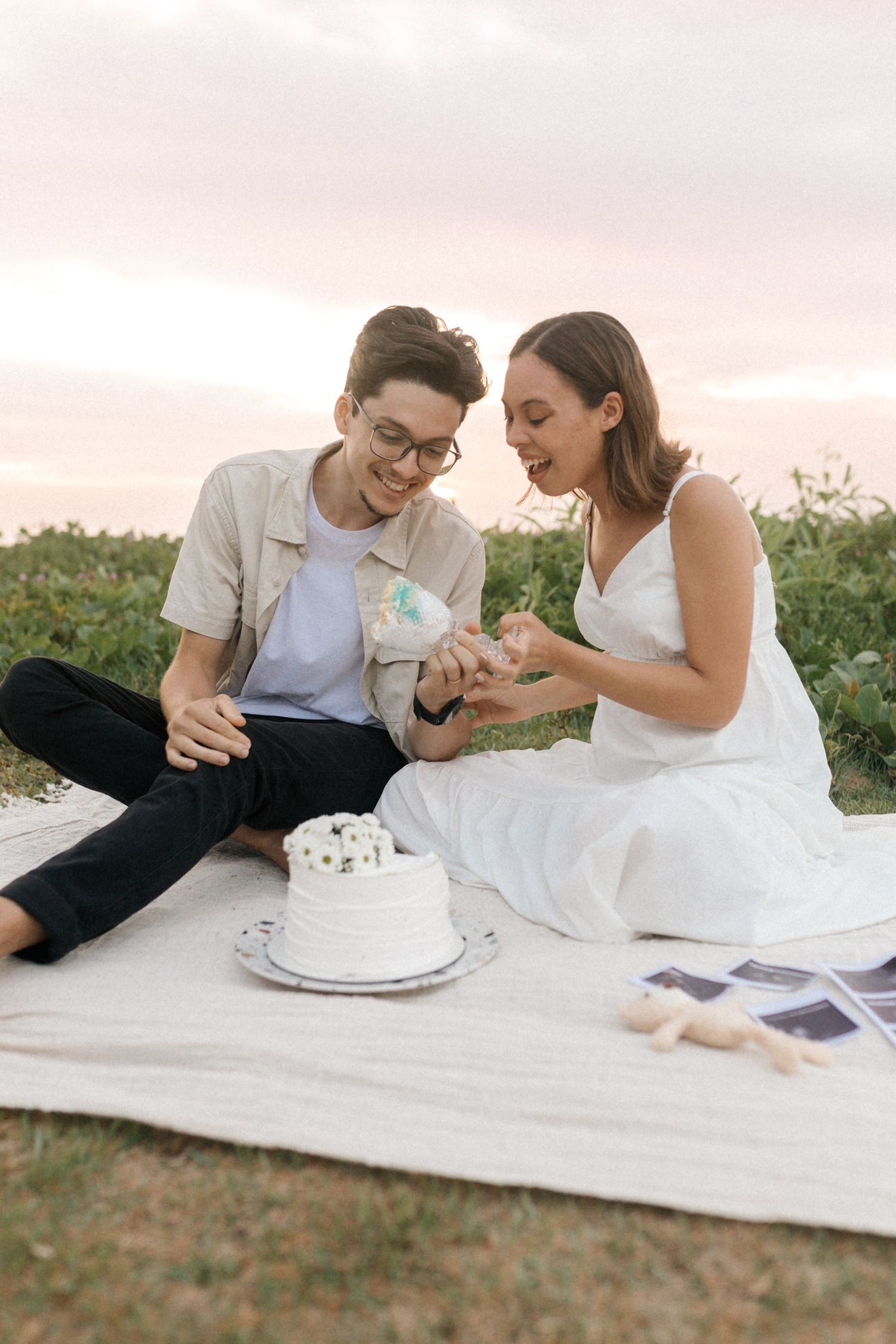 A couple sits on a blanket outdoors sharing cake and playfully feeding each other during a romantic picnic.