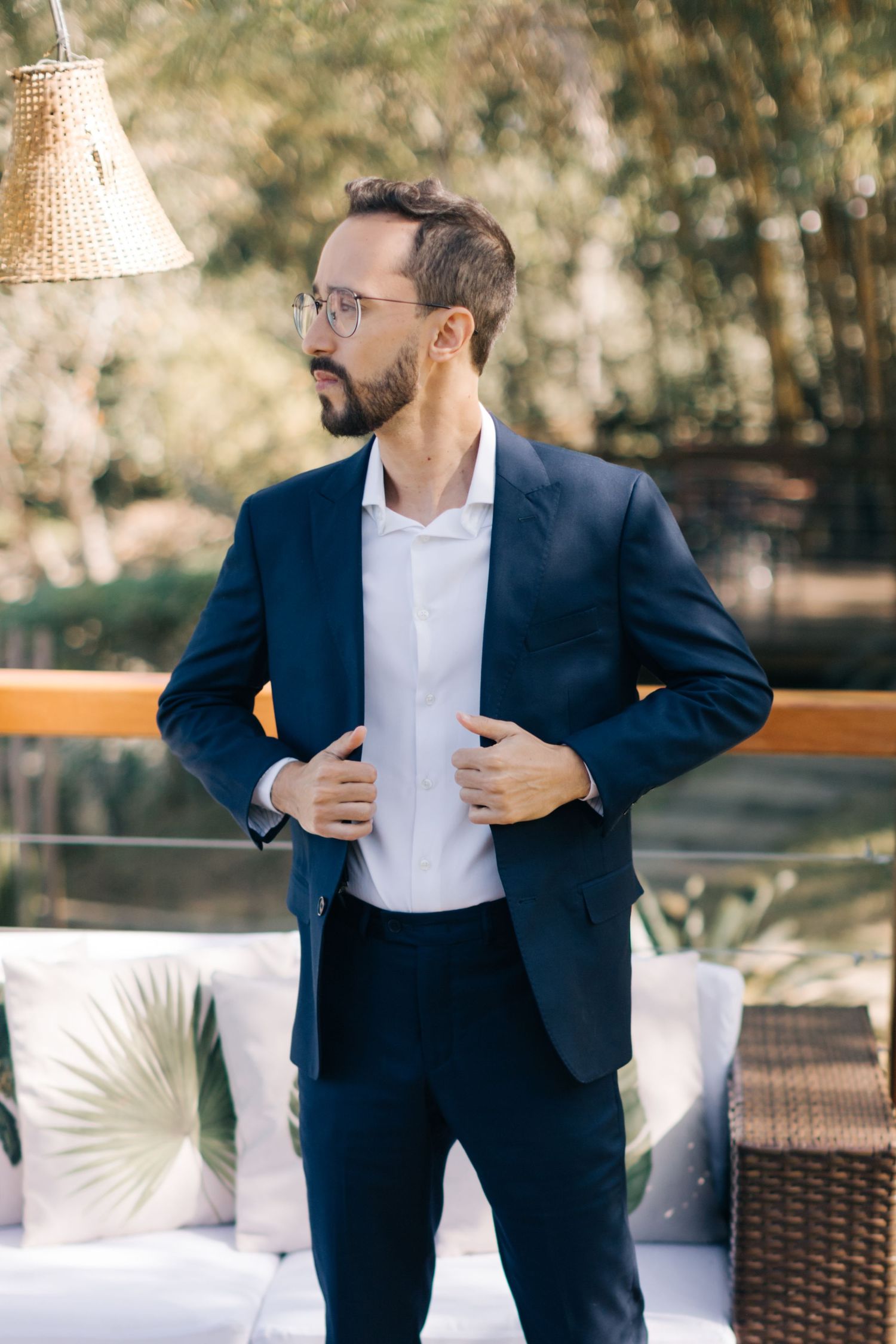 Groom in navy suit adjusting jacket on outdoor terrace with rattan furniture.