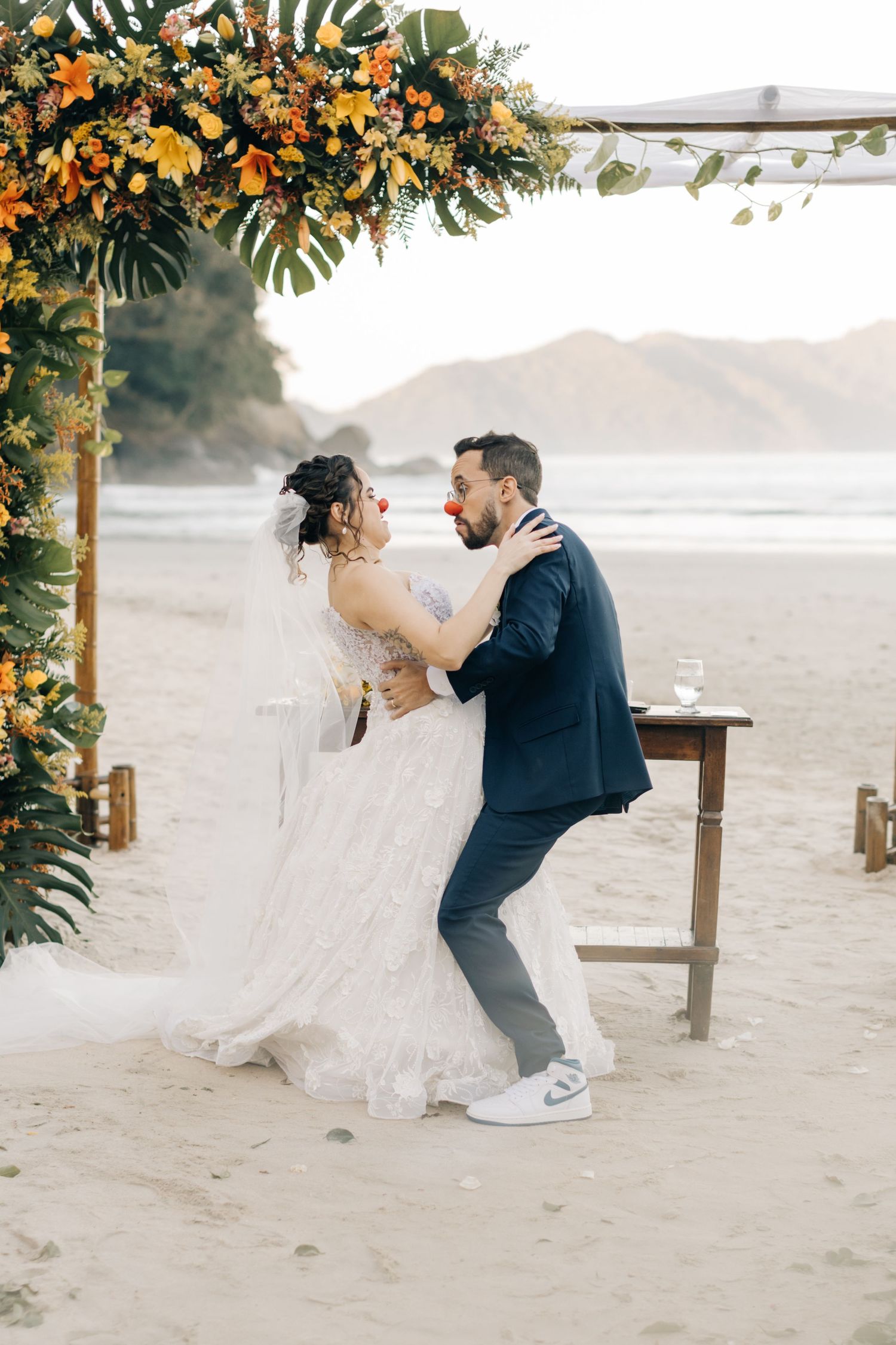 Beach wedding scene with couple dancing under tropical floral decorations and mountain backdrop.
