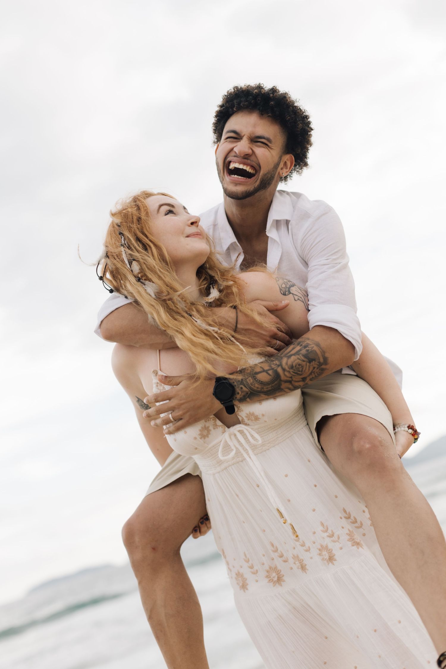 A couple in white clothing shares laughter and embraces during a seaside photoshoot.