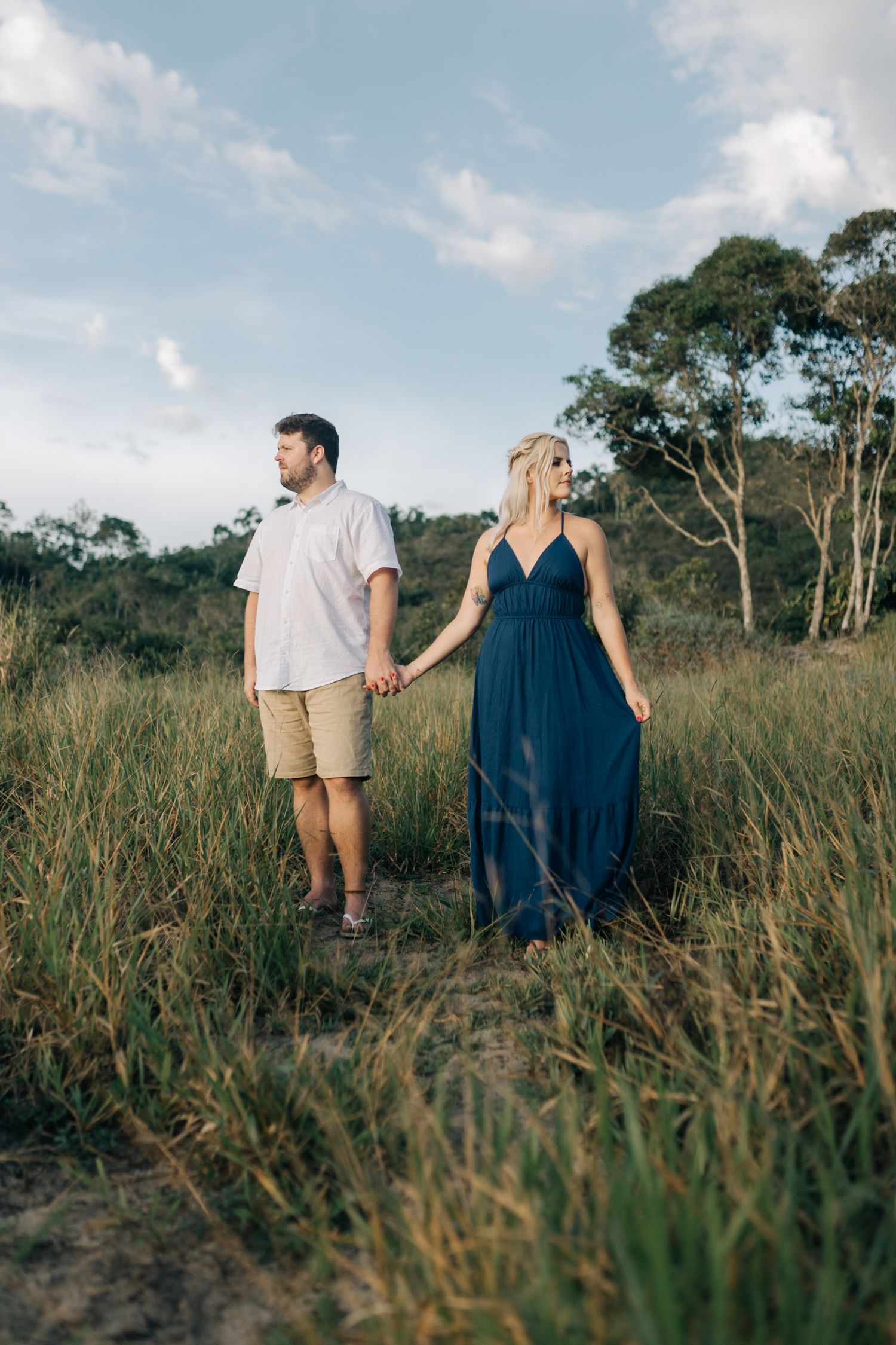 Walking couple holding hands through tall grass with mountains in background at dusk.