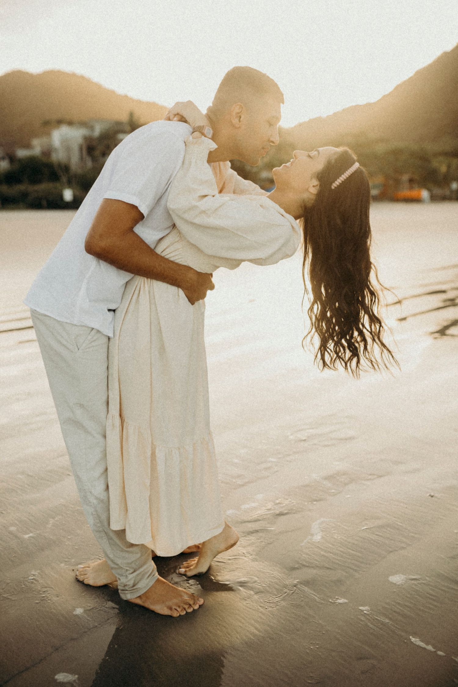 Couple in light colored clothing share romantic dip pose on beach at sunset.