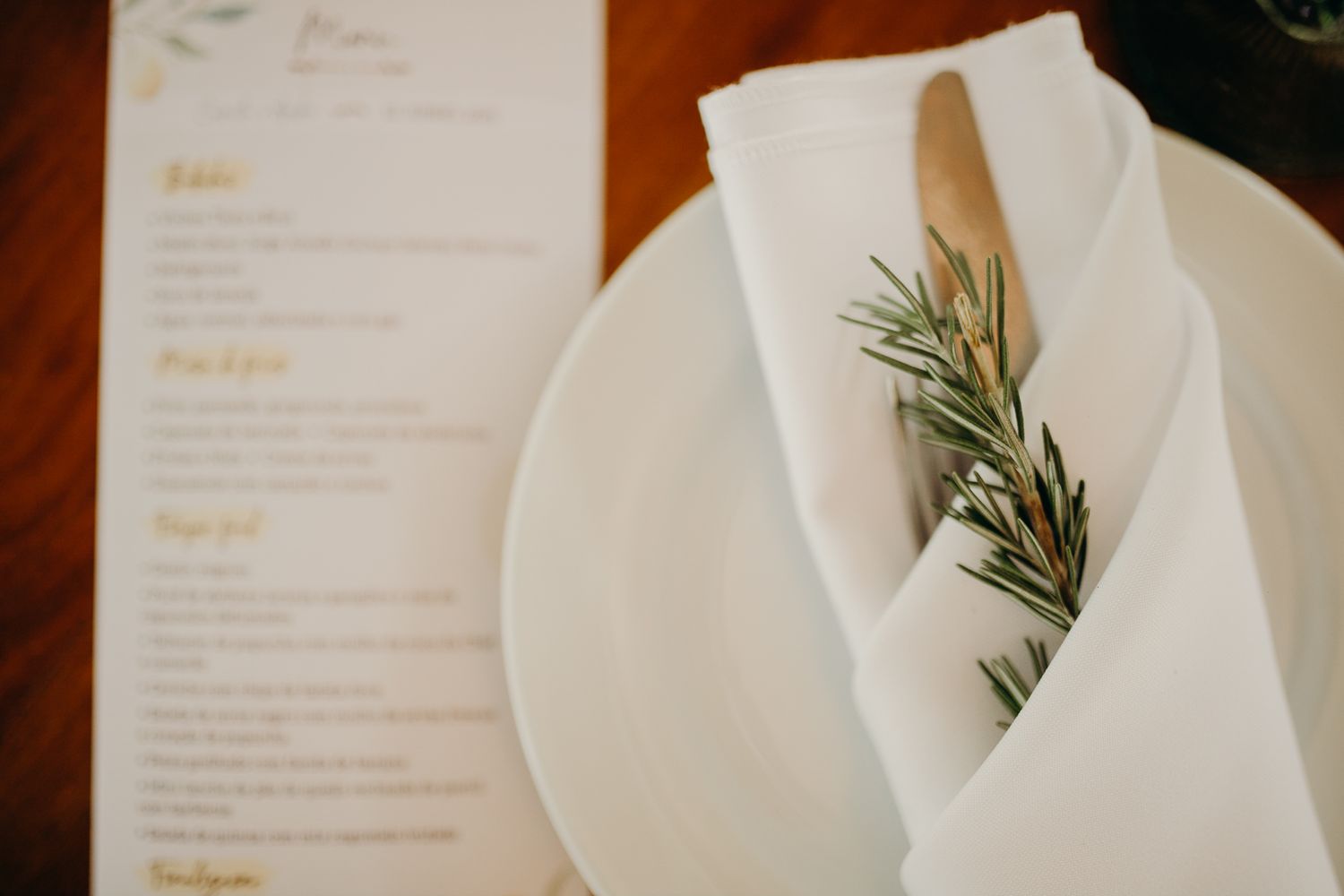 Close-up of white napkin with fresh rosemary sprig and menu card on dining plate.