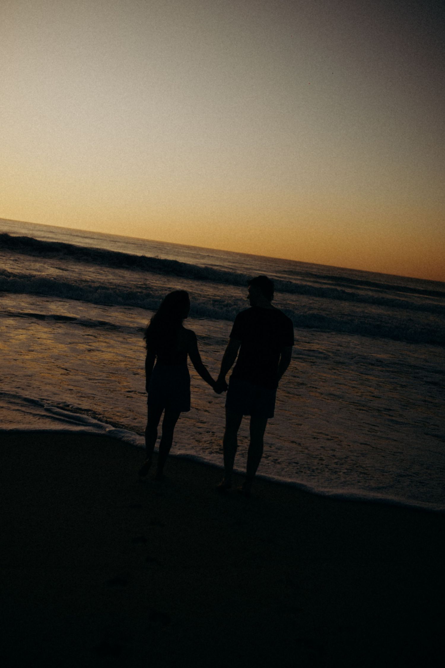 Silhouetted couple holds hands while standing on beach at sunset against orange sky and rolling waves.
