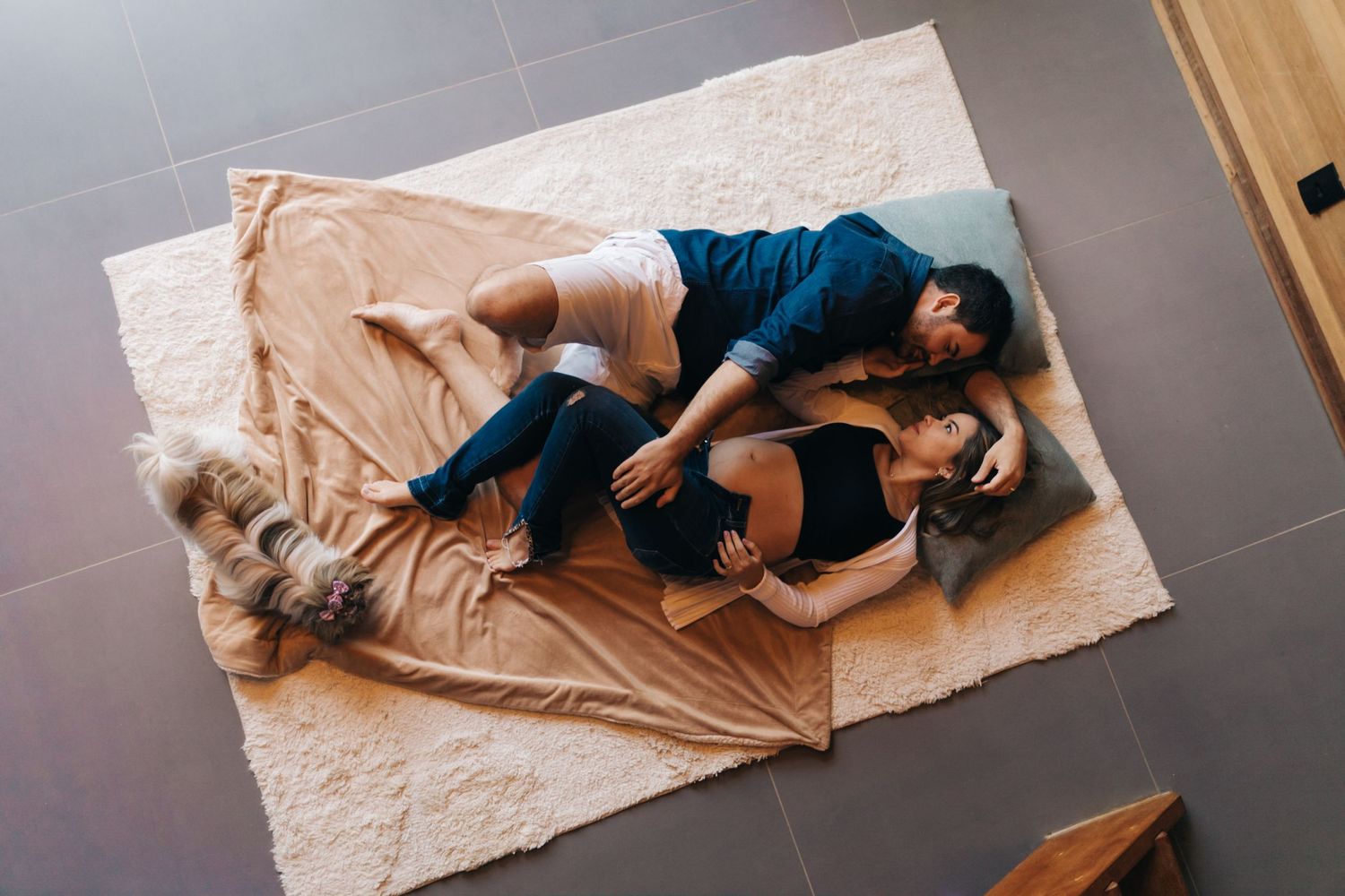 A couple lounges on a beige blanket with their dog on grey tile flooring.