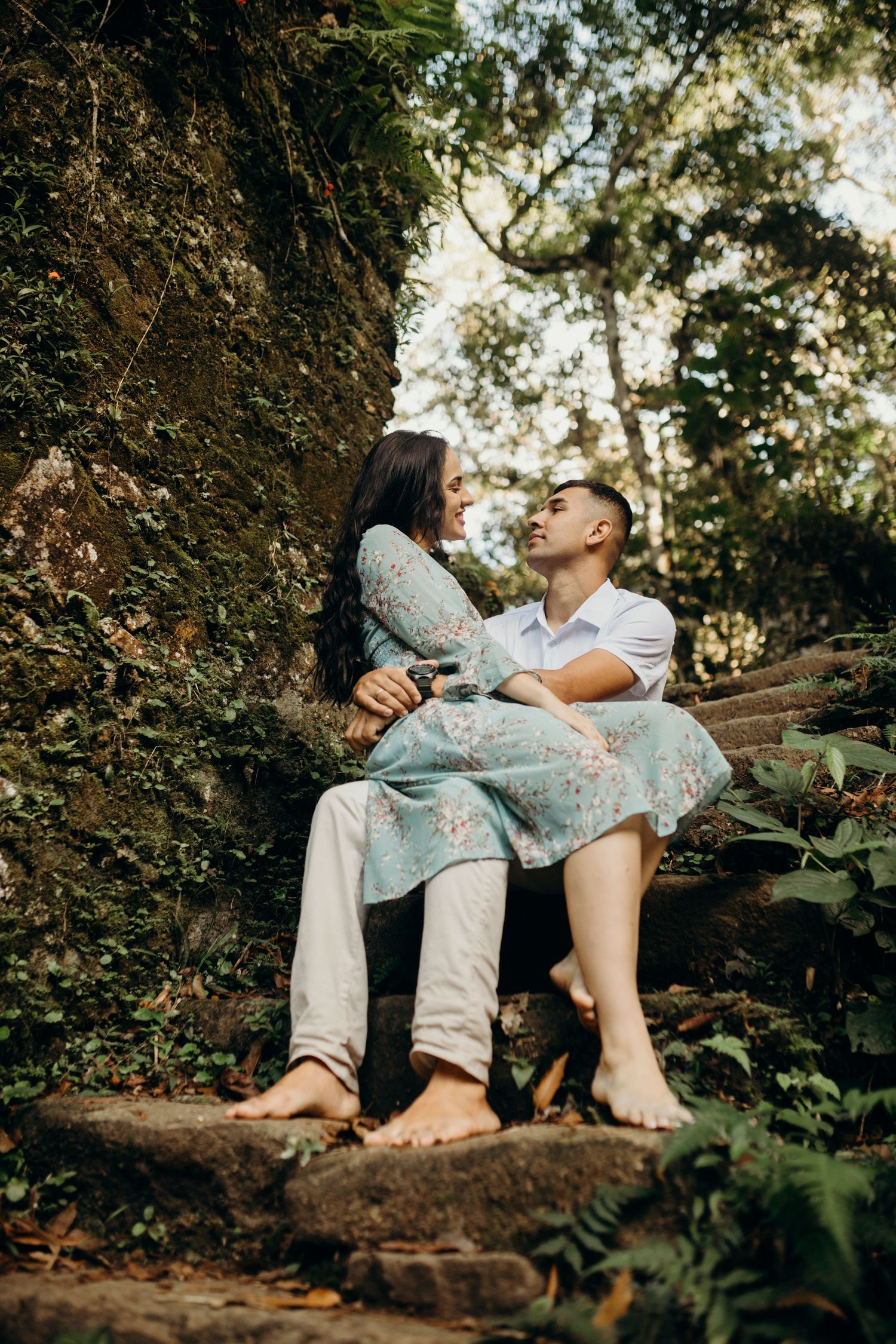 A couple sits barefoot on mossy steps sharing an intimate moment in nature.