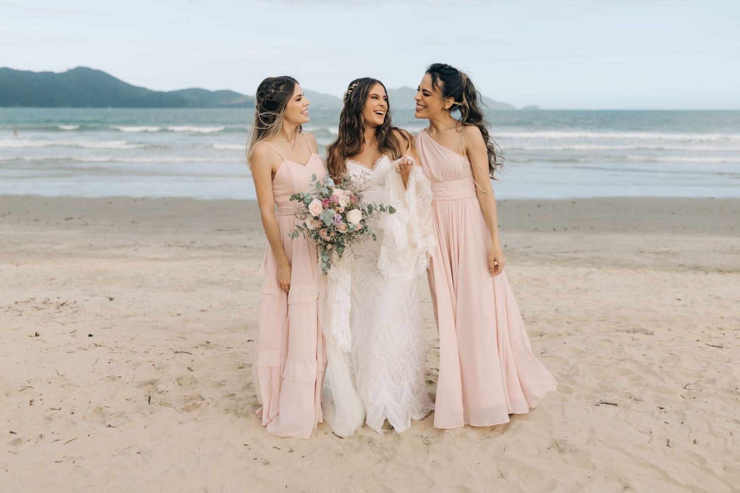 Bridal party in pink and white dresses pose together on sandy beach with ocean backdrop.