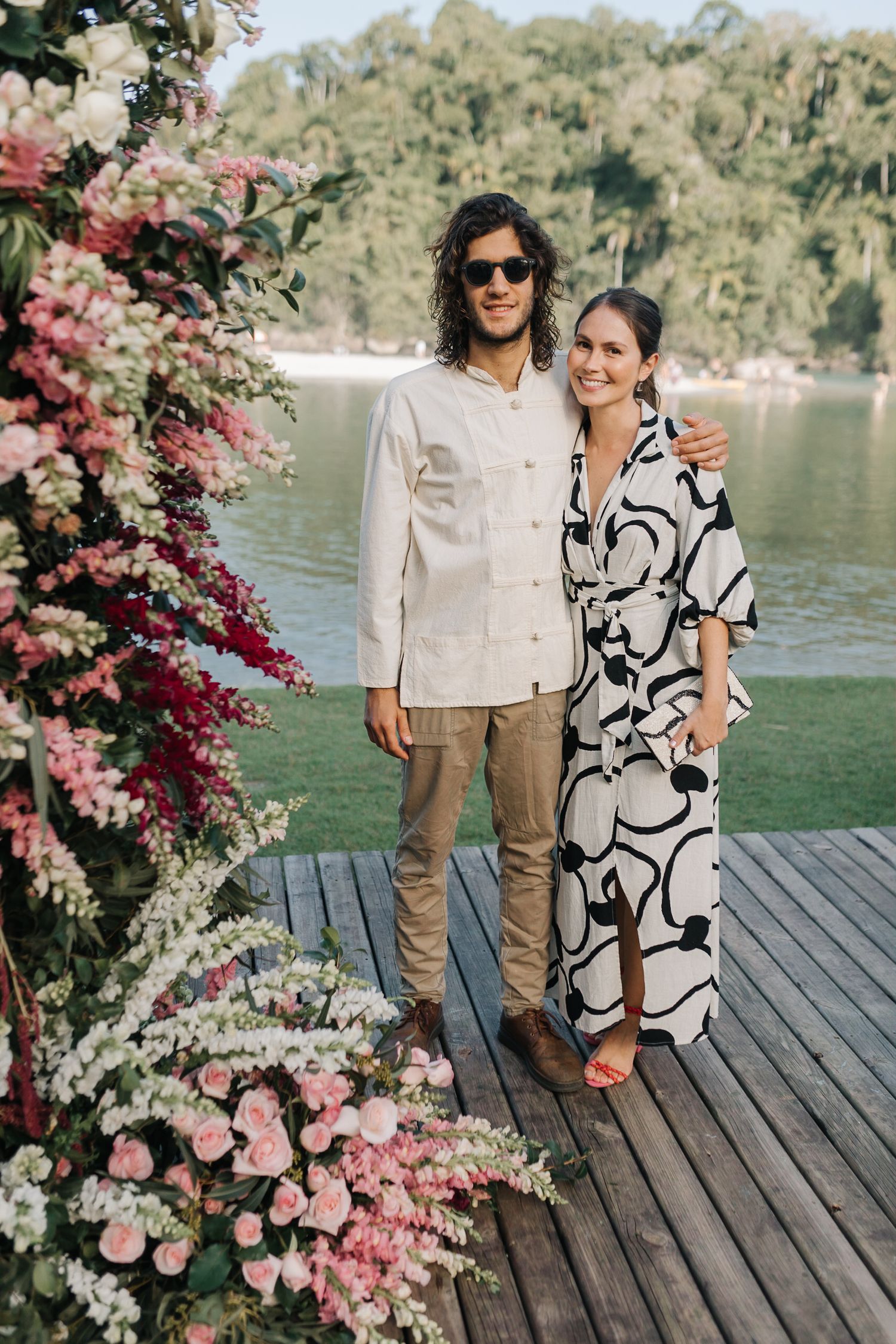 Couple poses on wooden deck decorated with pink floral arrangements by the water.