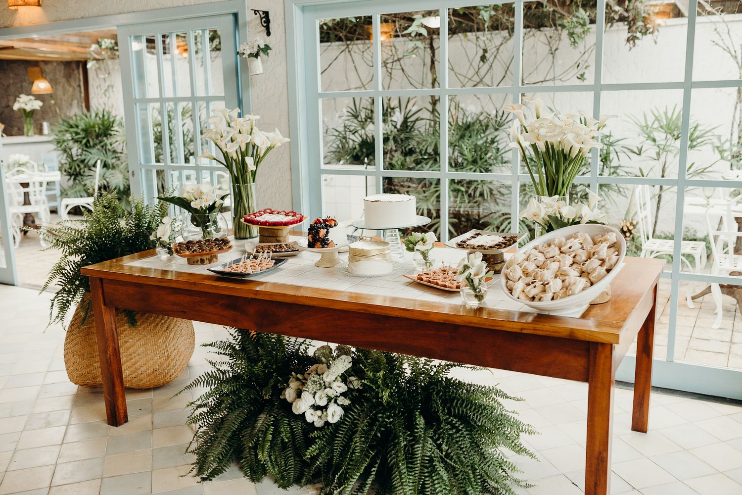 Long wooden banquet table decorated with greenery and white flowers in an indoor-outdoor space.