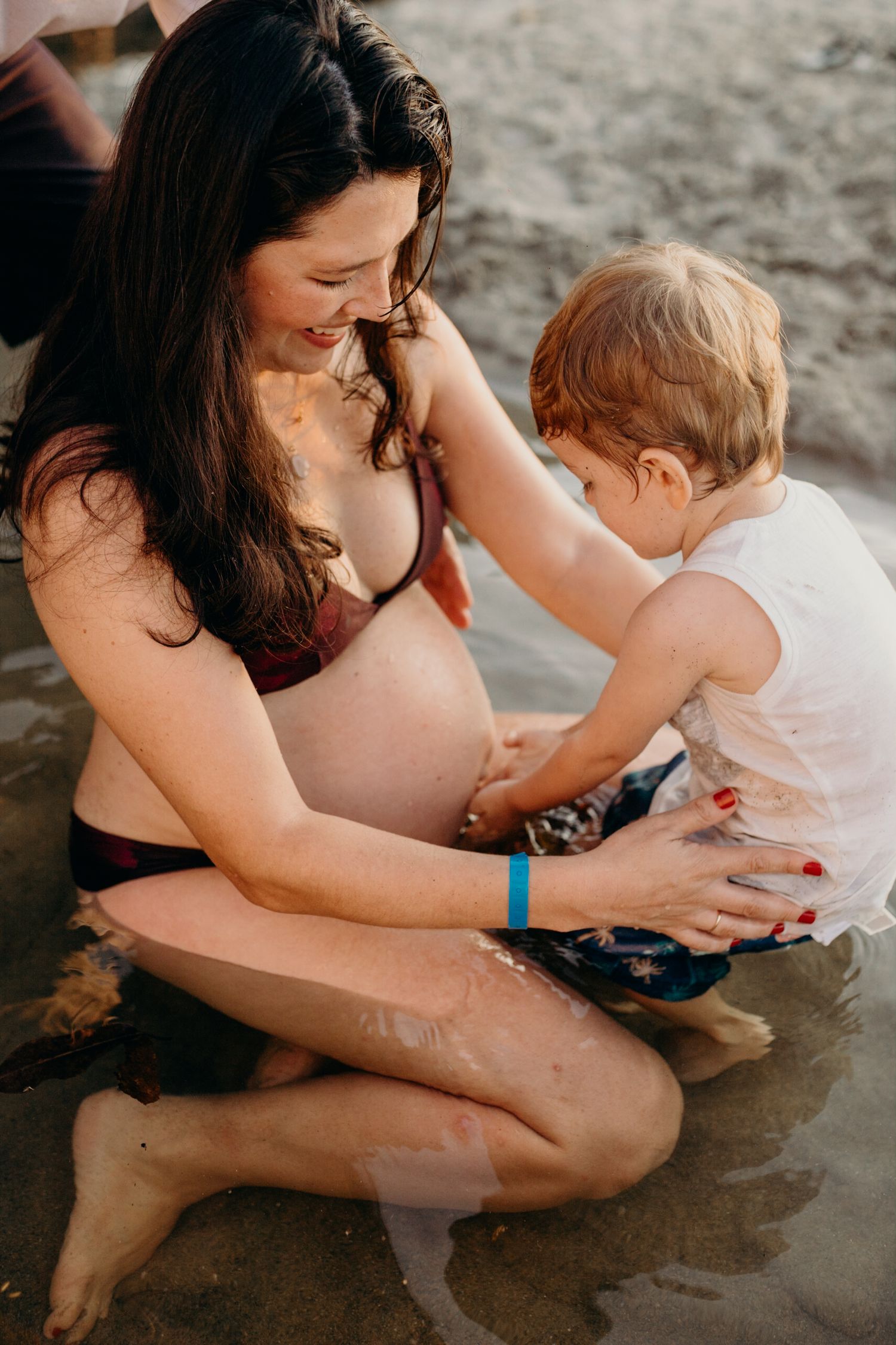 A pregnant woman in a black swimsuit plays with a toddler in shallow beach water during a sunny day.