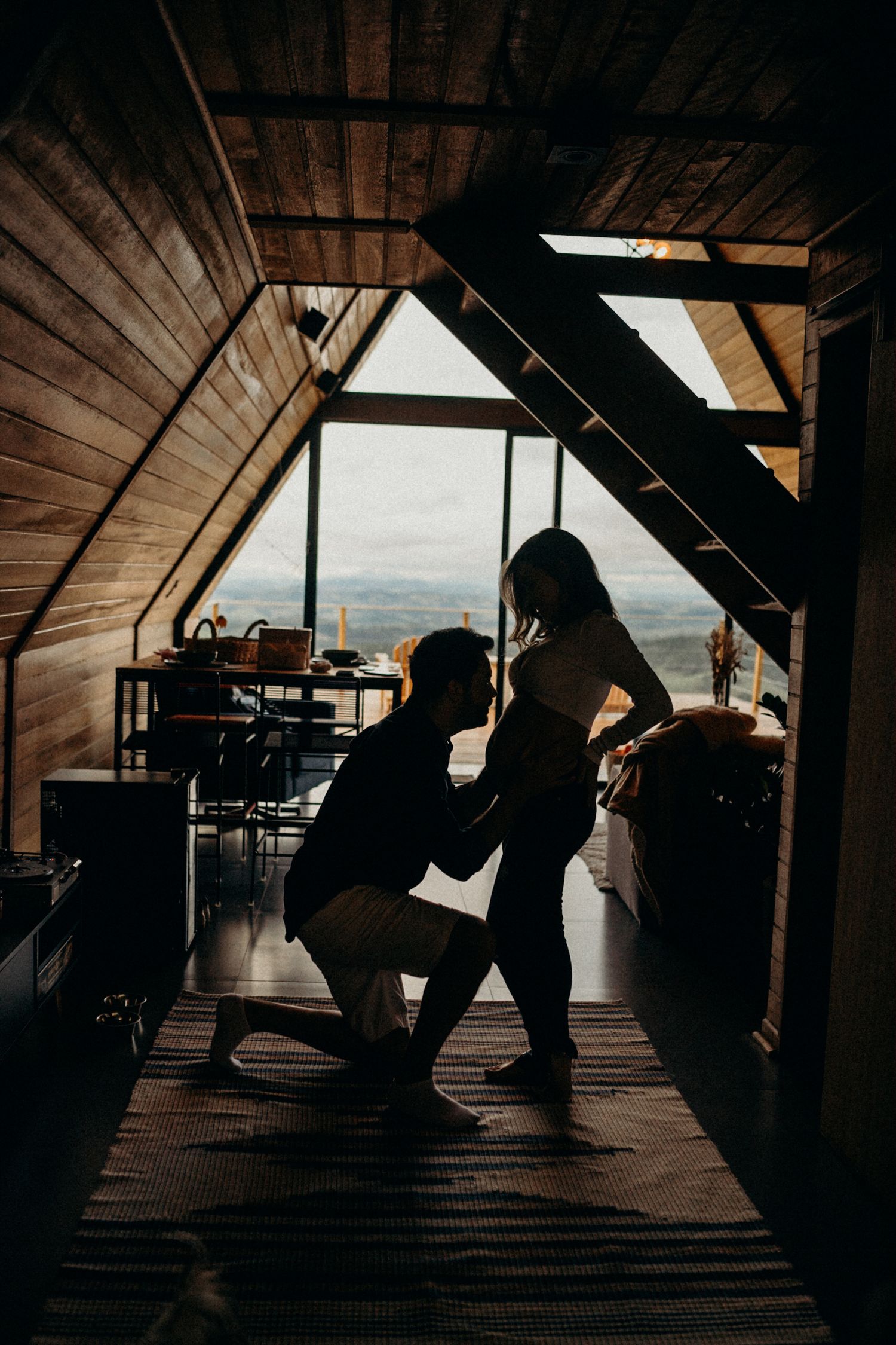 A silhouetted proposal moment in an A-frame cabin with mountain views visible through the windows.