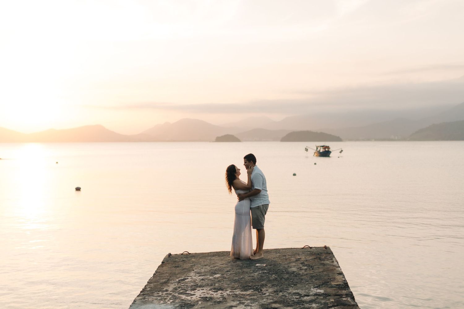 A romantic couple embraces on a pier at sunset overlooking a tranquil lake with mountains in the background.
