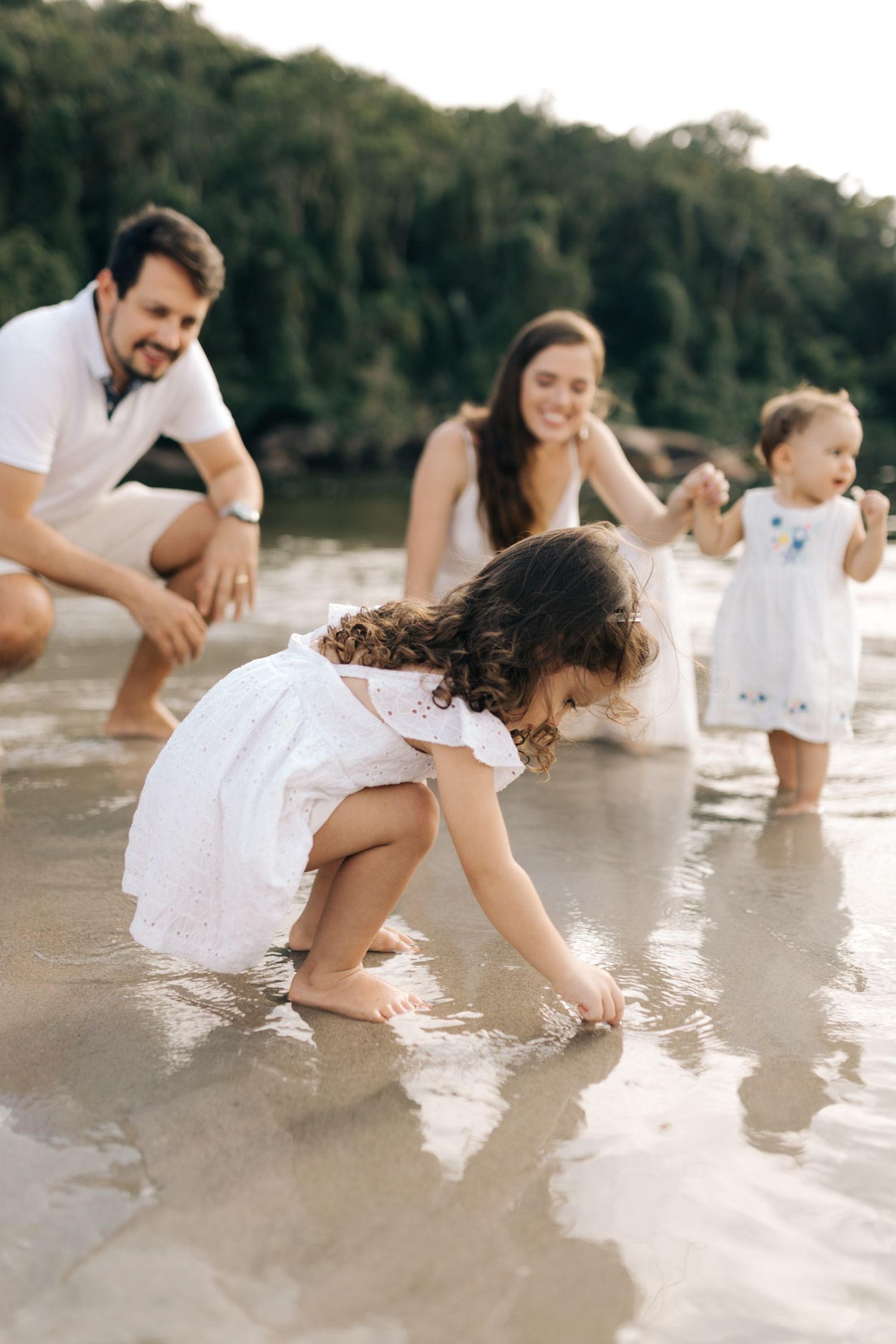 Family watching child explore in shallow water during beach photoshoot.