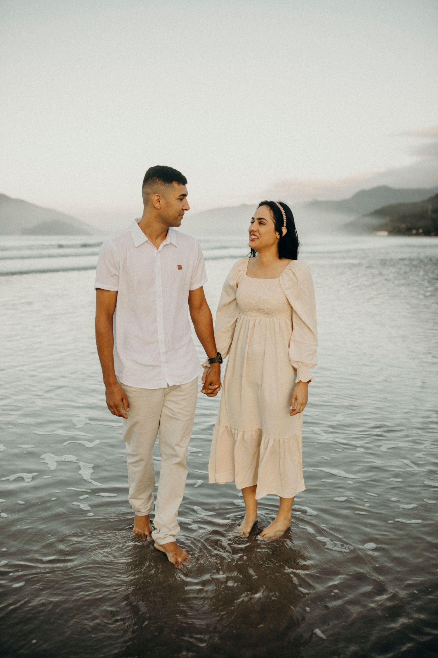 Couple standing in shallow ocean water wearing light colored outfits against mountain backdrop.
