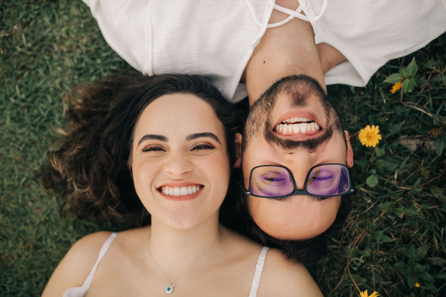 Couple laying together on grass with yellow flowers wearing white outfits during an outdoor photoshoot.
