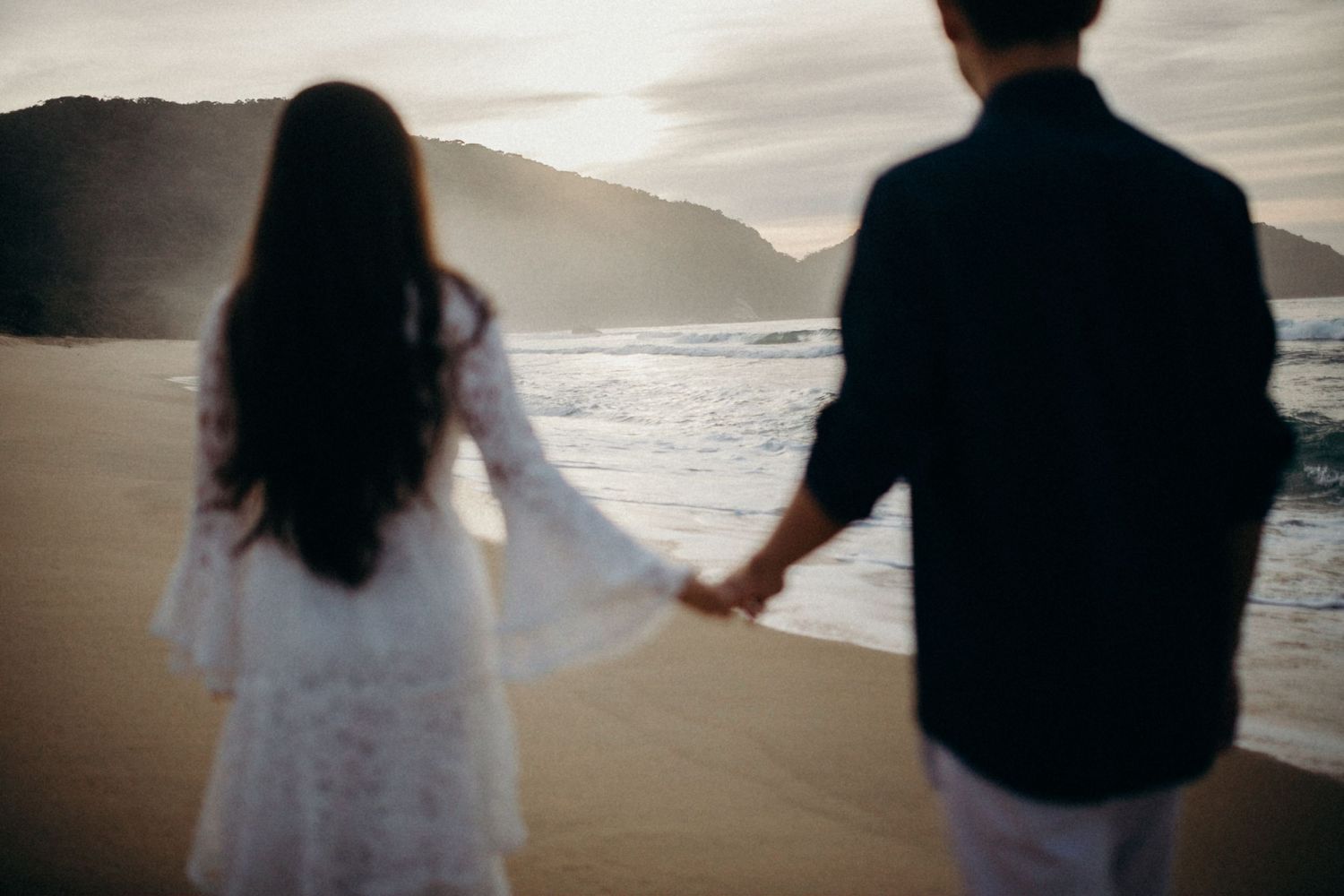 Back view of couple holding hands while walking along a misty beach with mountains.