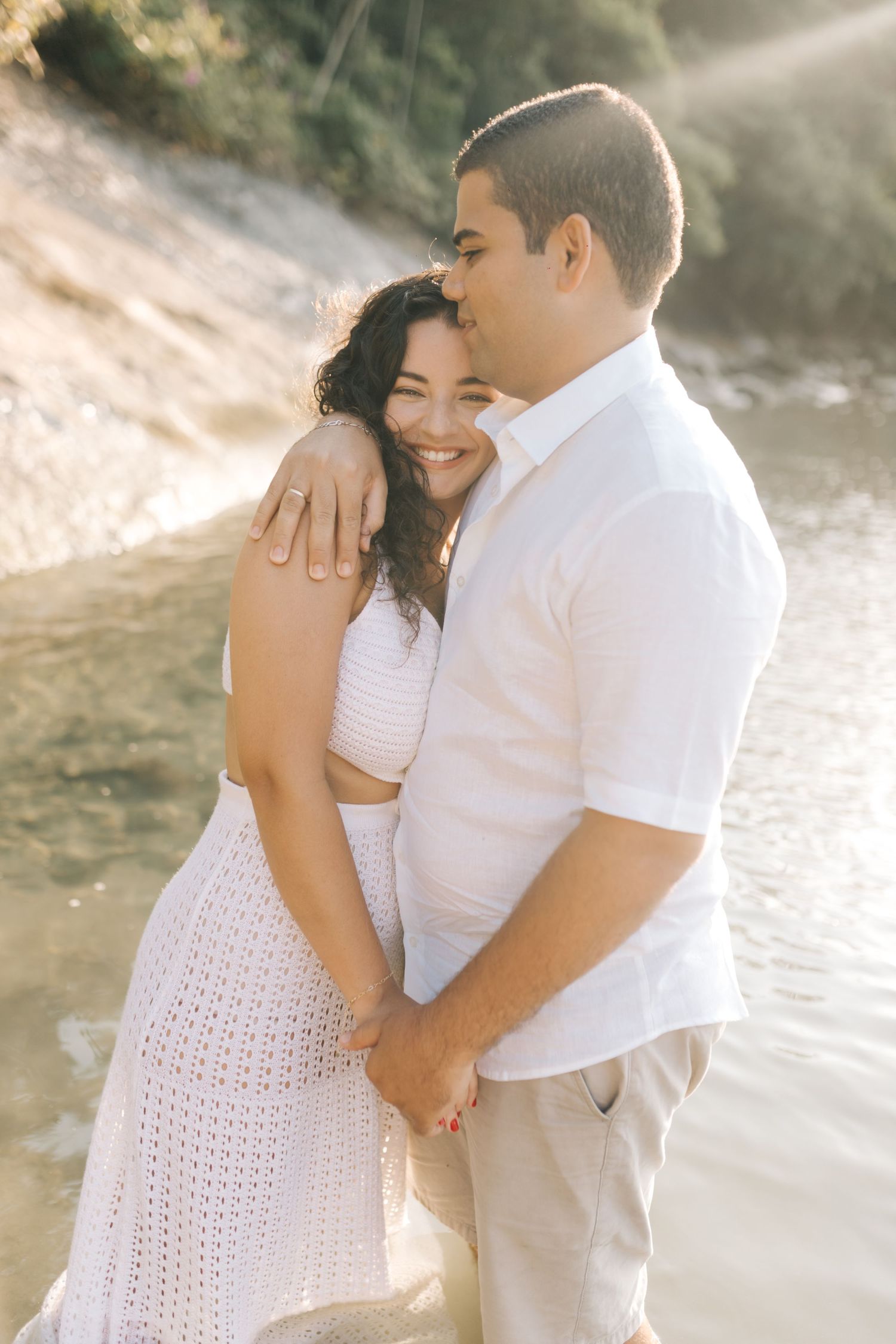Couple sharing intimate embrace on beach wearing white and flowing attire in sunlit setting.