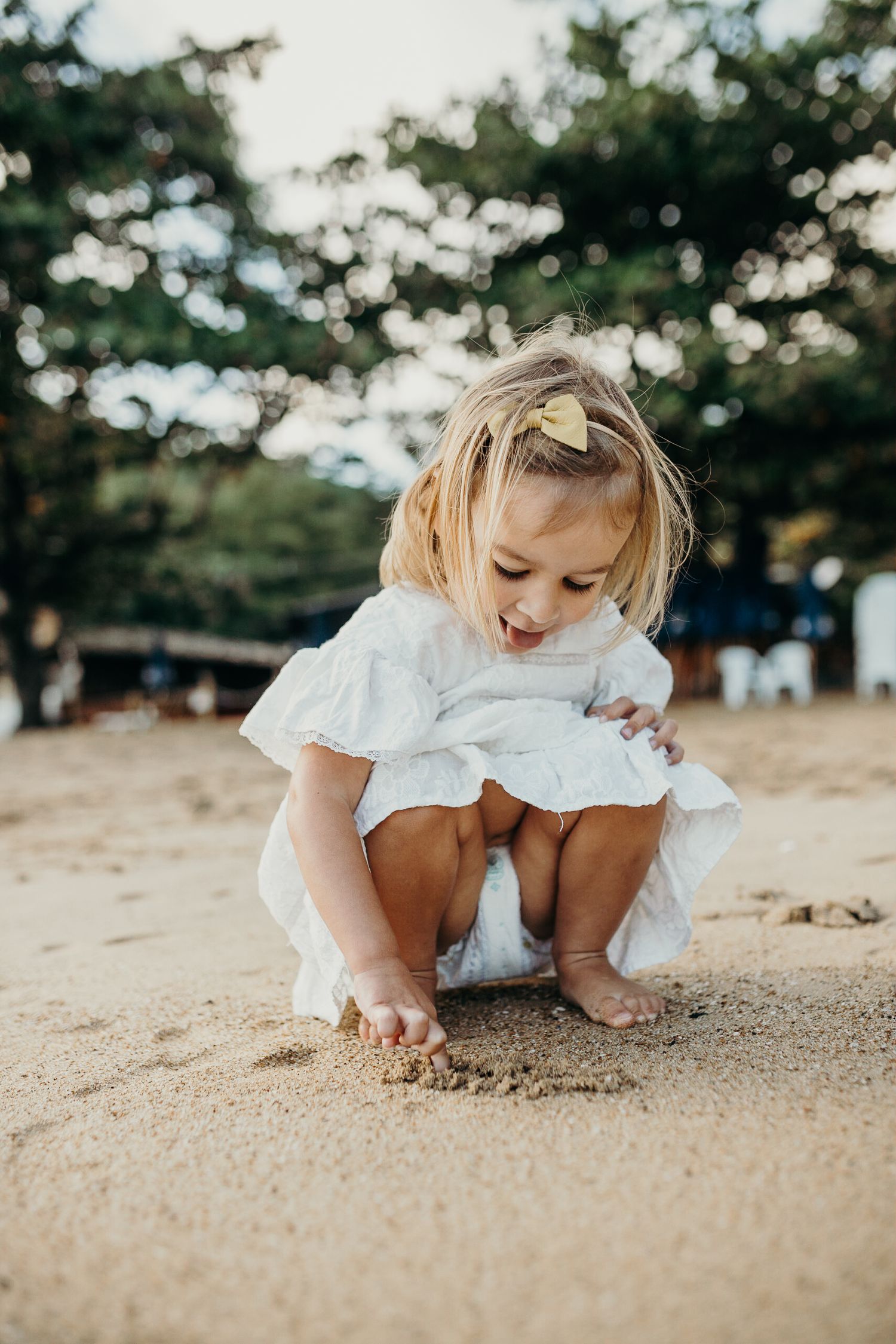 A child in white dress crouches down to explore the sandy beach.