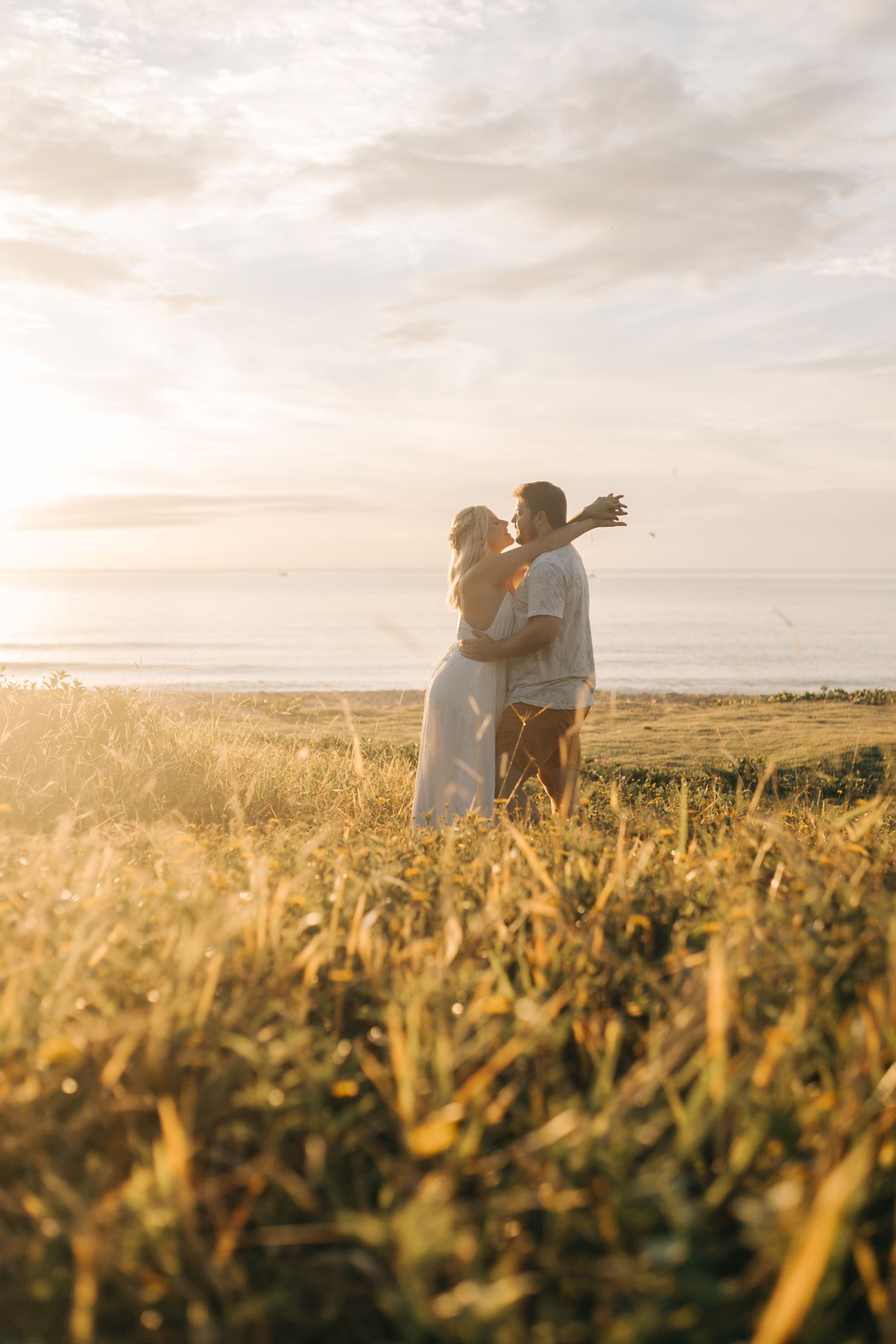 A romantic couple embraces in a sunlit field at golden hour with tall grass swaying in the breeze.
