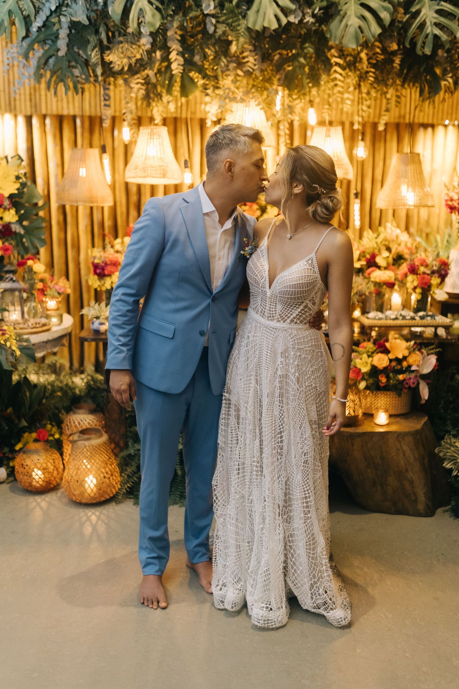 Wedding couple in blue suit and lace dress posing against bamboo wall with floral decorations.