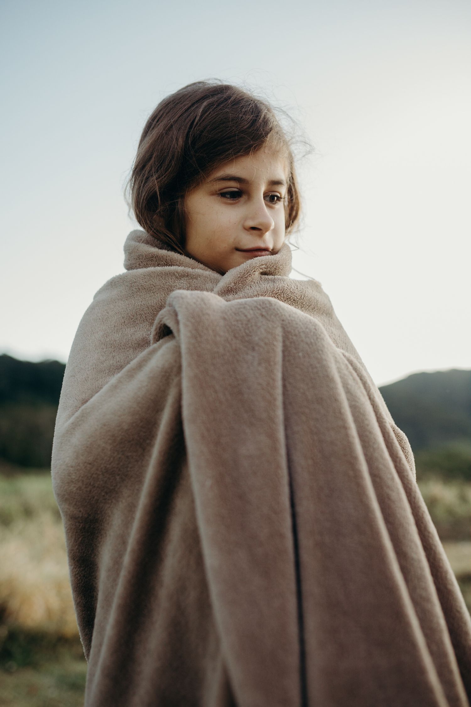 Person wrapped in a soft beige blanket looking over shoulder in outdoor setting.