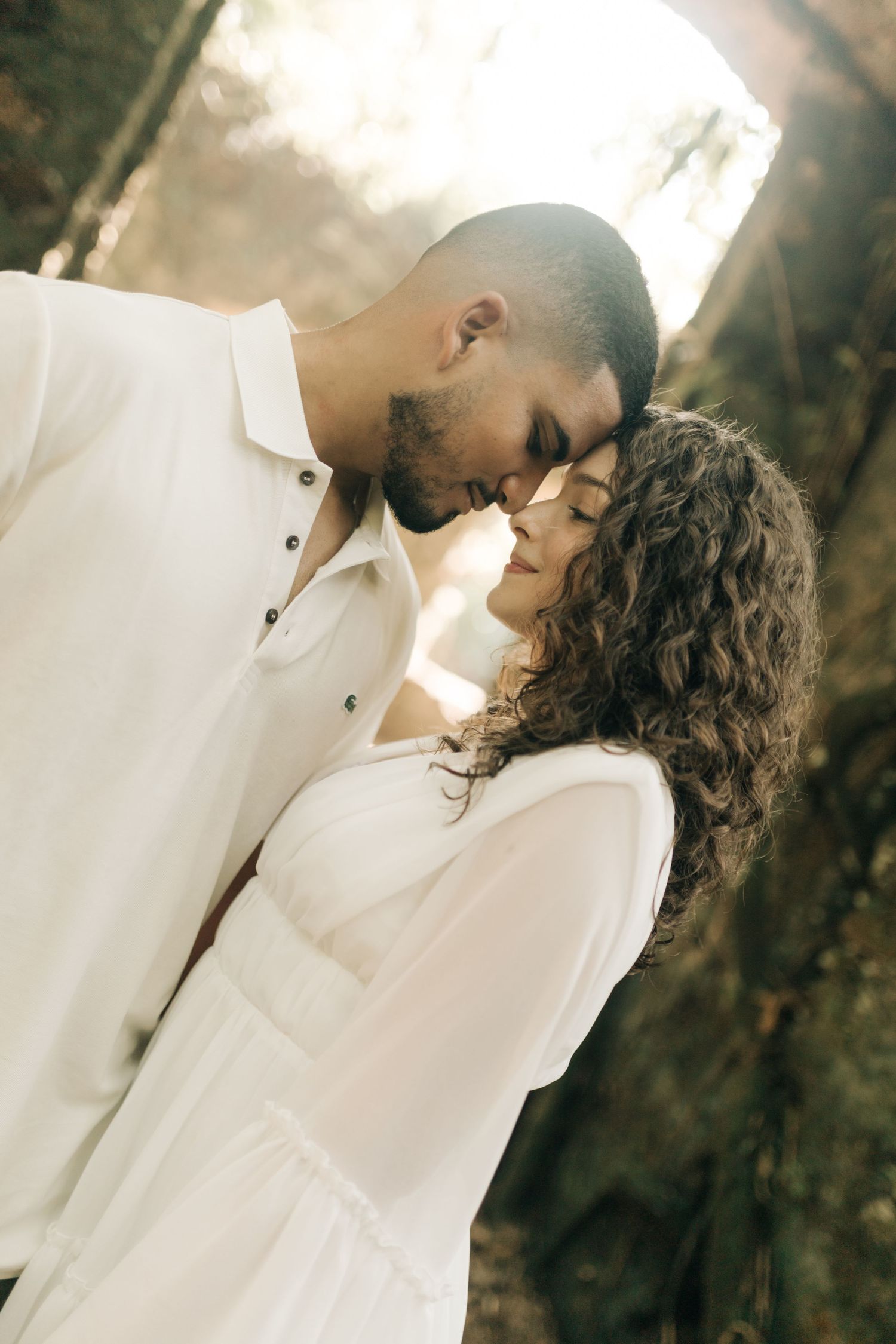 Dreamy sunlit portrait of couple in white sharing intimate moment in wooded setting.