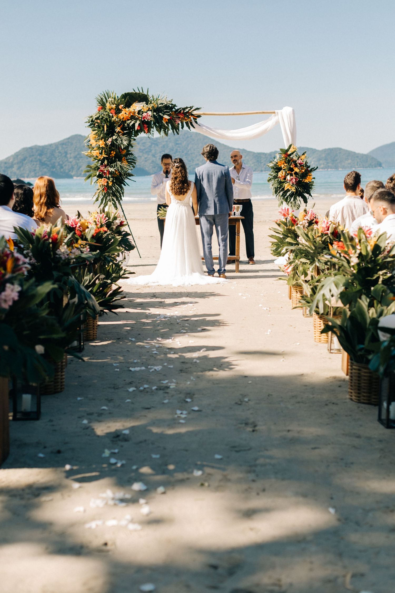 A beachside wedding ceremony setup with tropical floral arch and mountain backdrop.