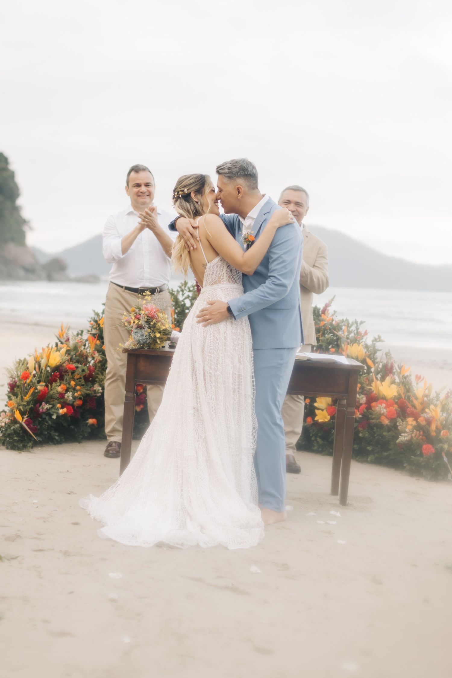 A romantic beach wedding ceremony scene with a couple in a white dress and light blue suit against an ocean backdrop.