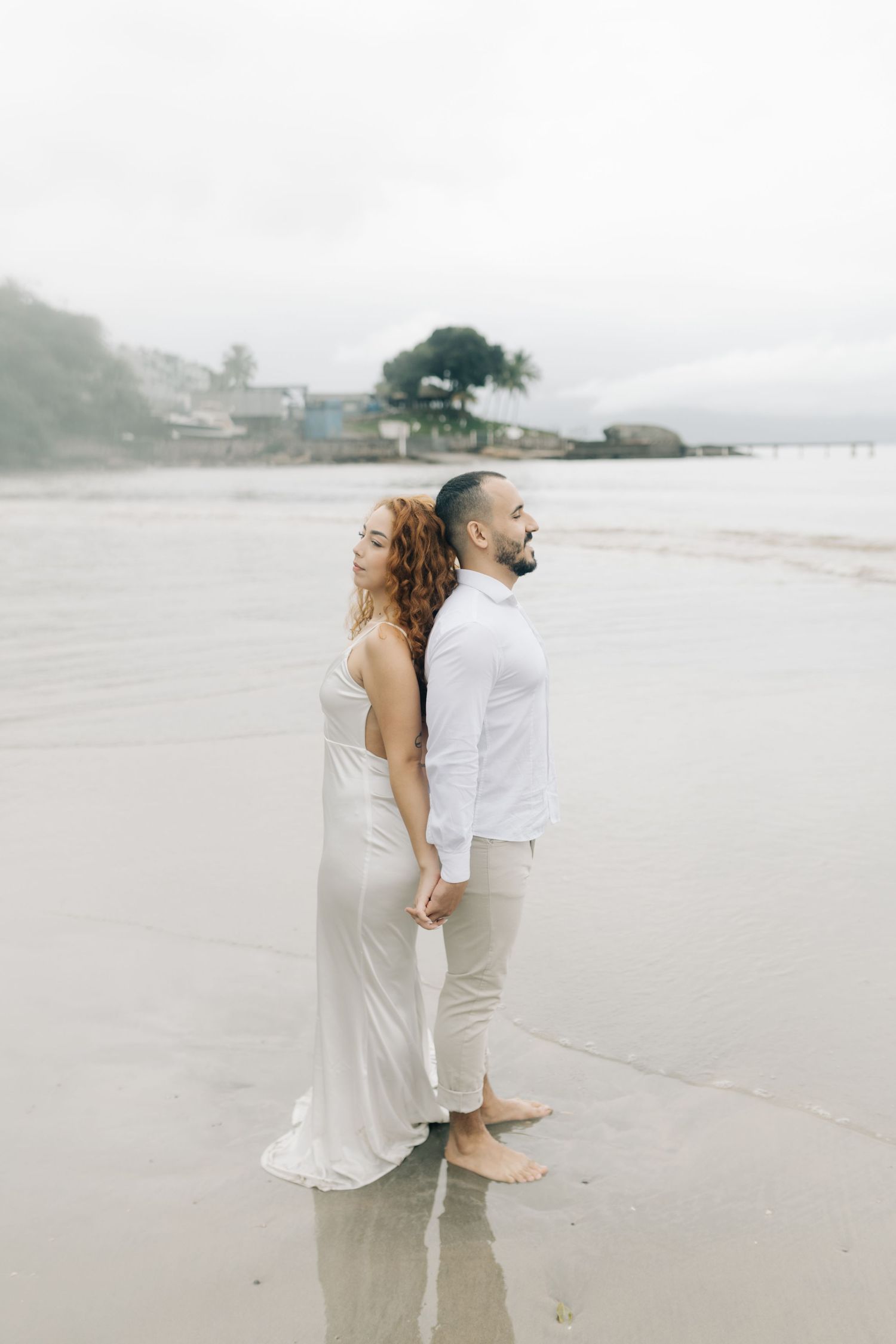 Couple stands together on a foggy beach holding hands in white outfits.