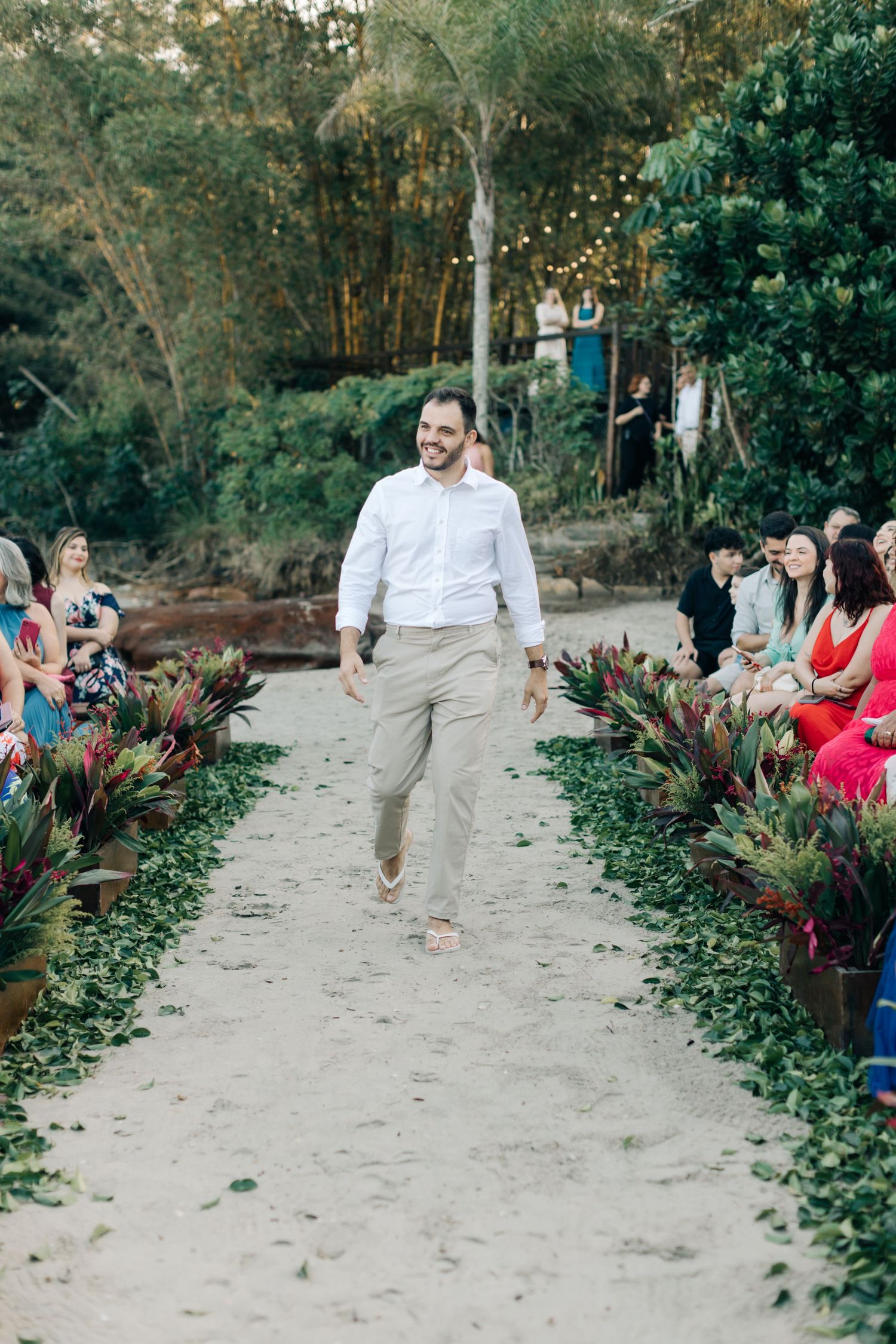 Wedding party member walks sandy aisle between tropical plants at outdoor ceremony.