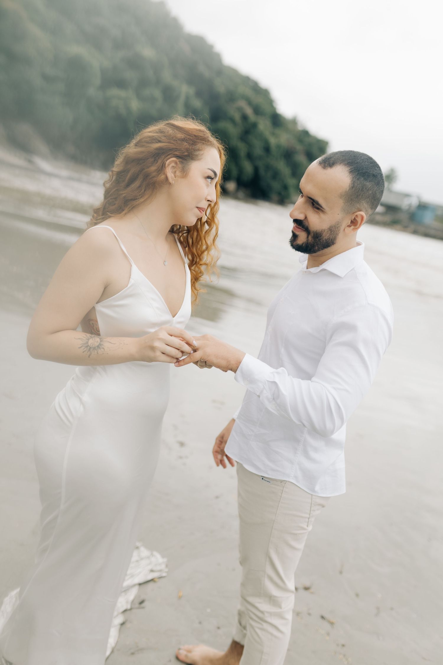 Couple in white clothing shares tender beach moment during engagement.