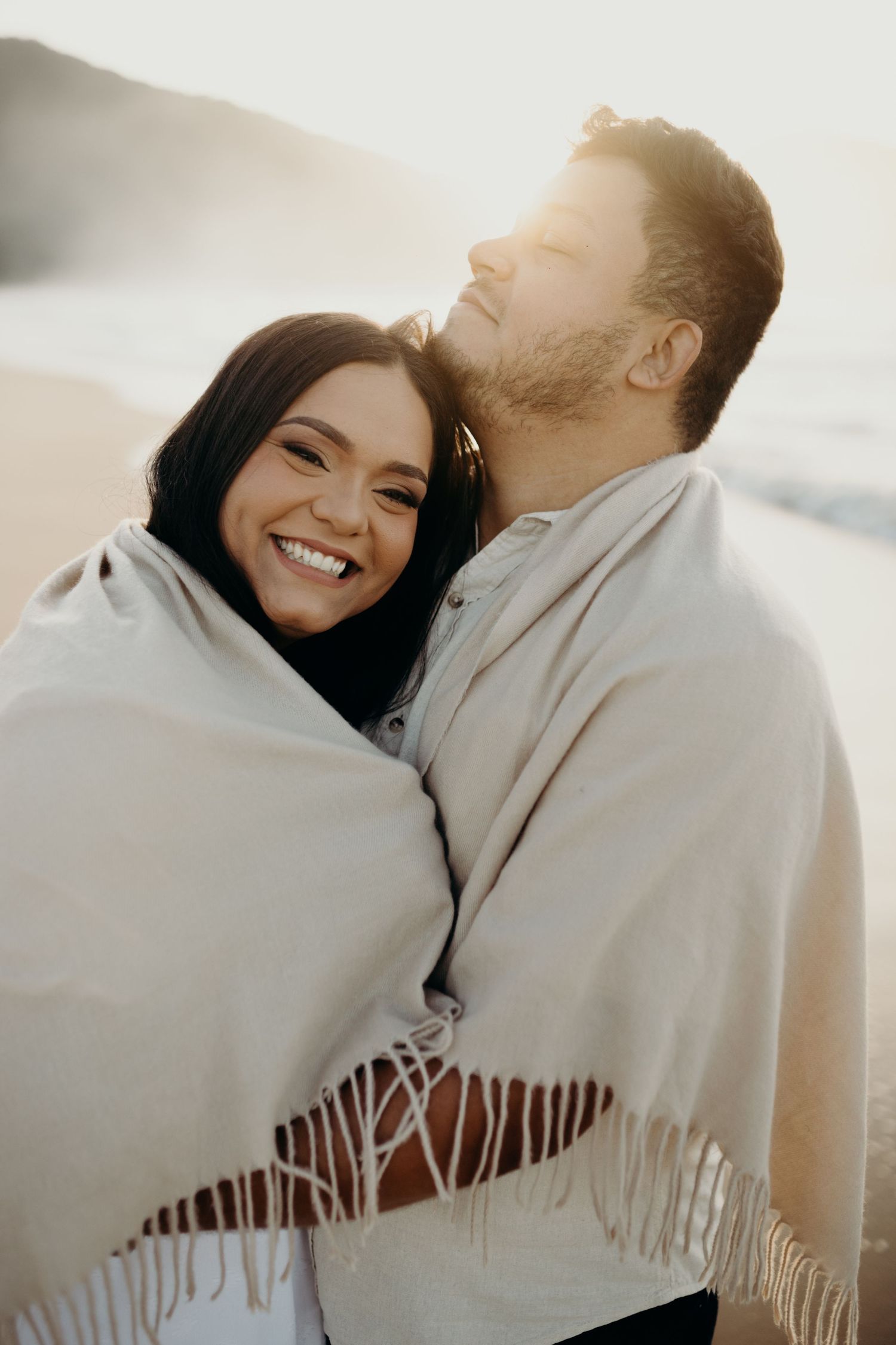 Joyful couple wrapped in white shawl sharing laughs during beach photoshoot.