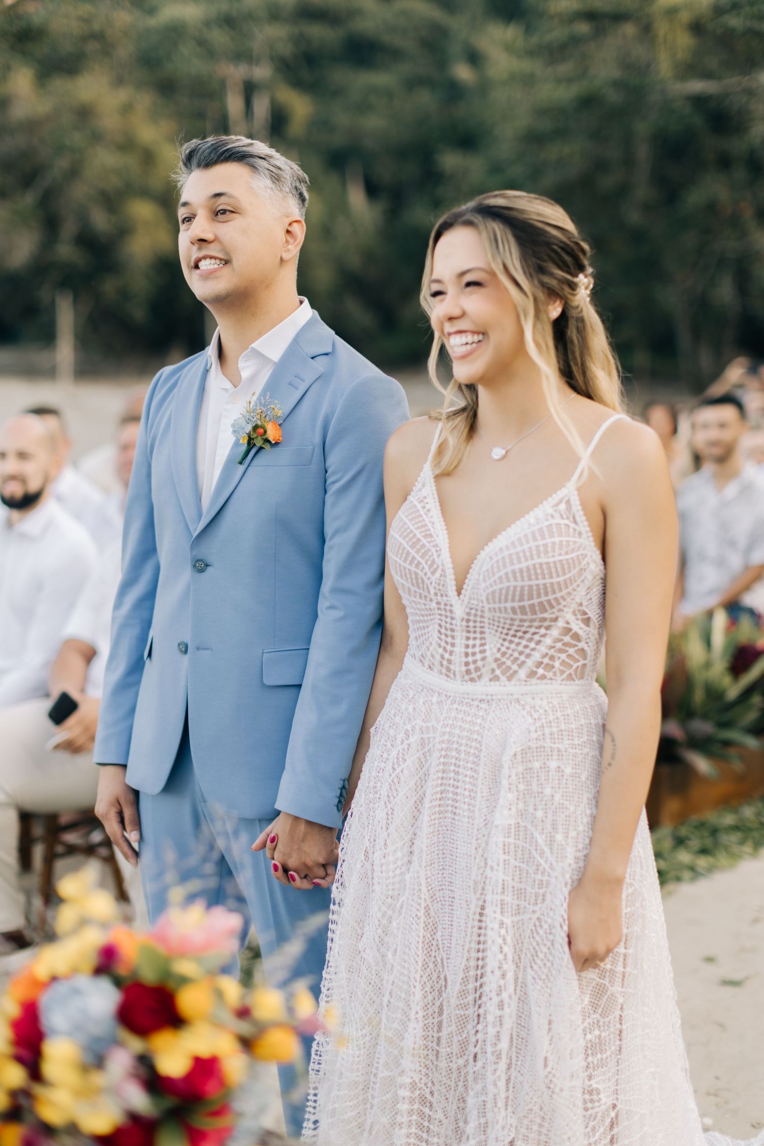 Beach wedding ceremony with light blue suit and white lace dress against ocean backdrop.