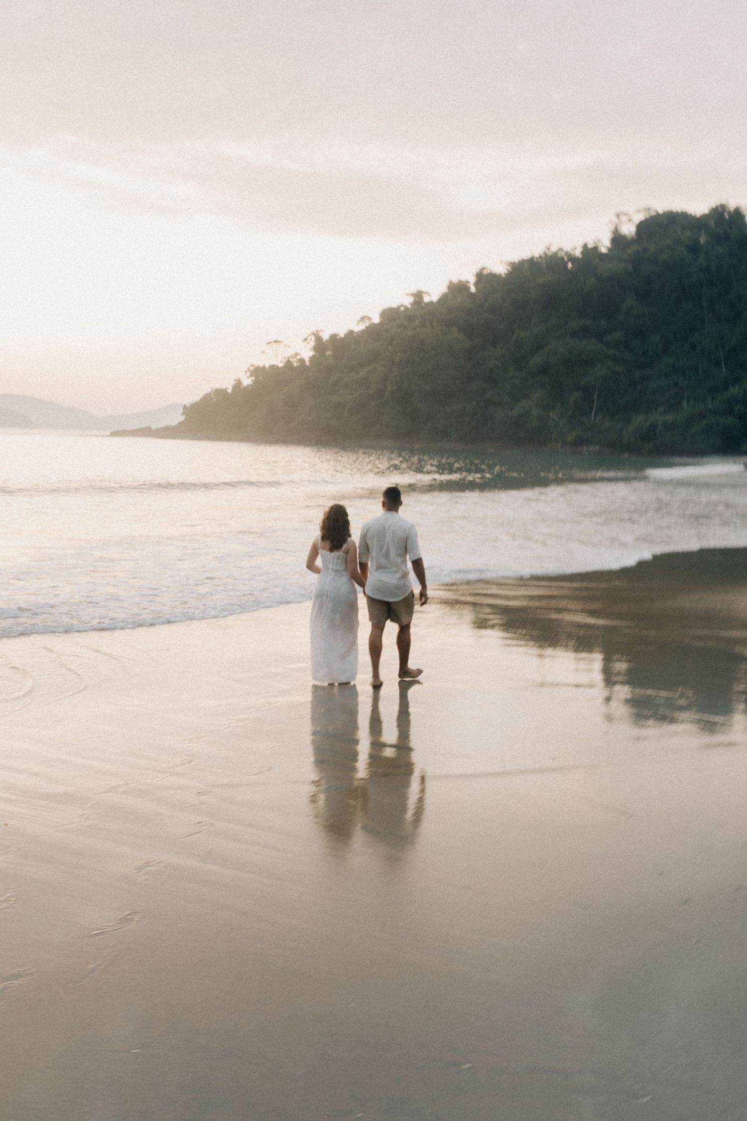 Couple walking hand in hand along a scenic beach at sunset with mountains in the distance.