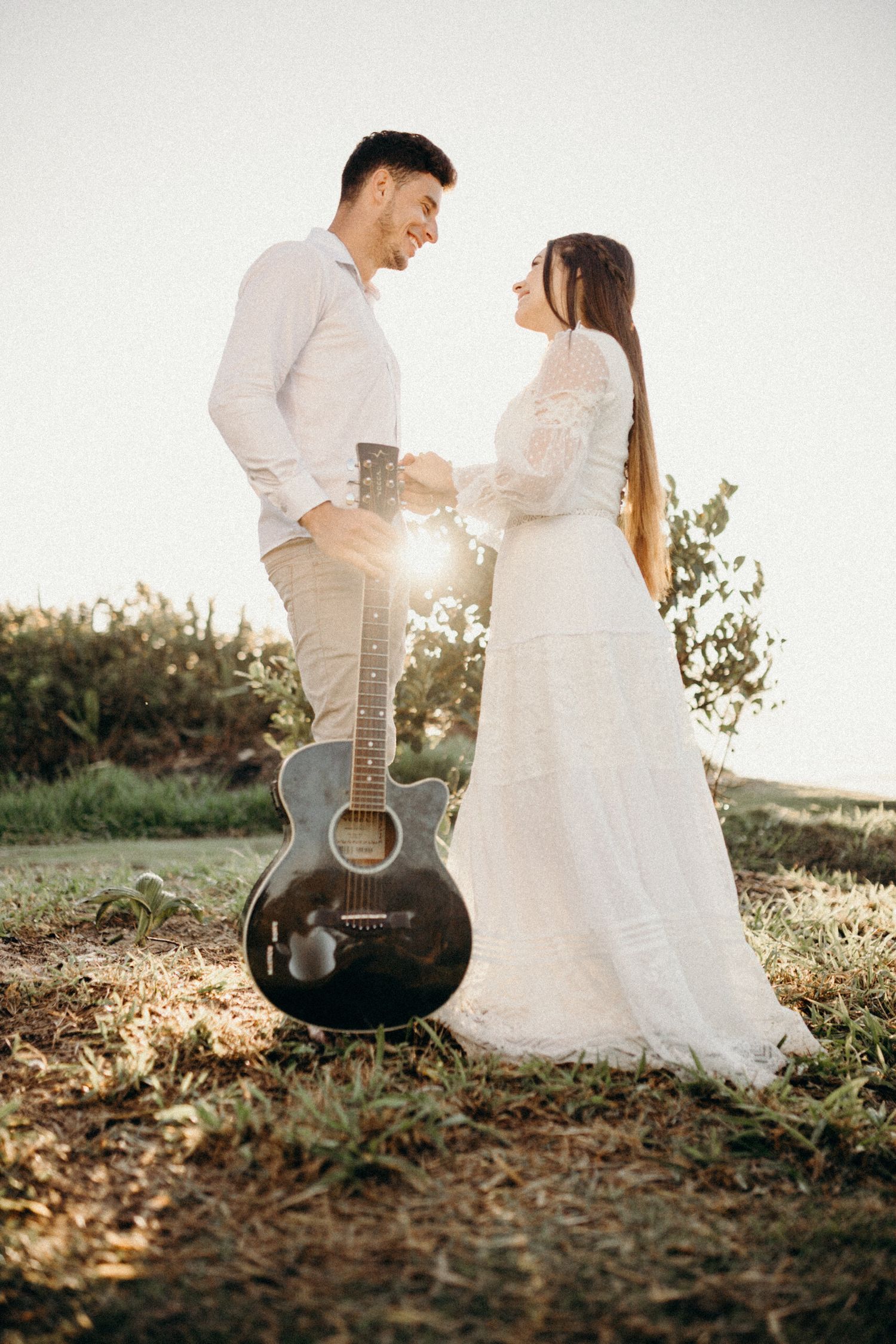 Romantic couple in wedding attire pose with black acoustic guitar in natural outdoor setting.