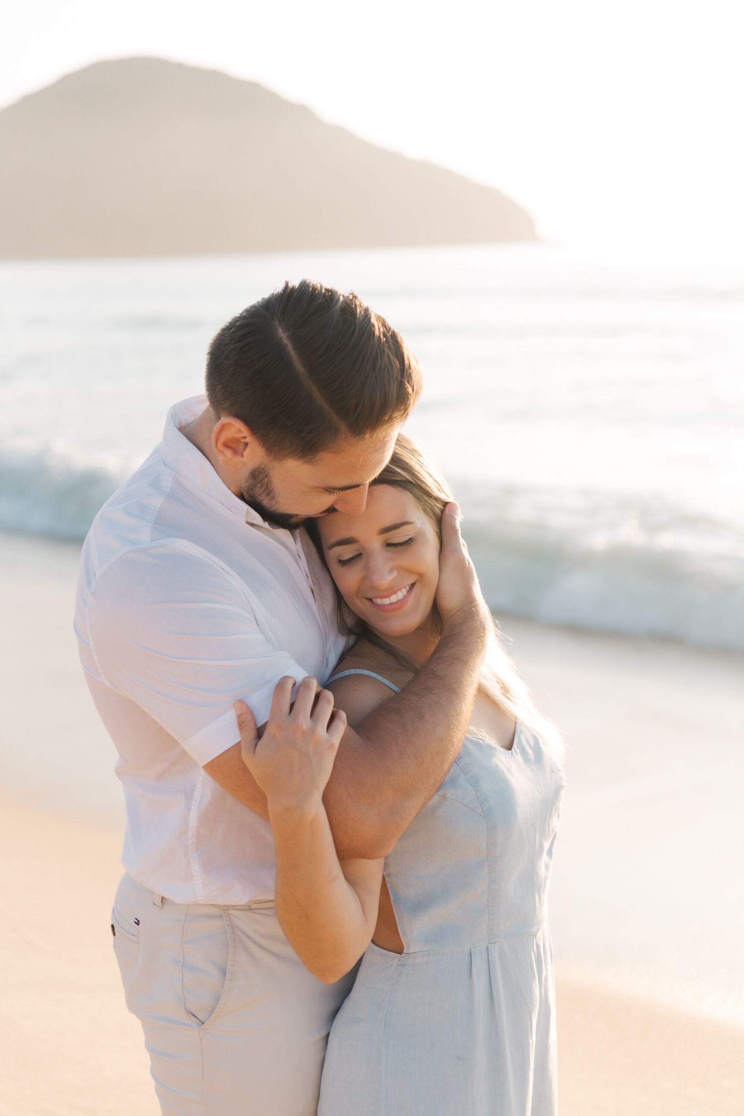 Loving couple sharing intimate moment on beach with ocean waves and mountains in background.