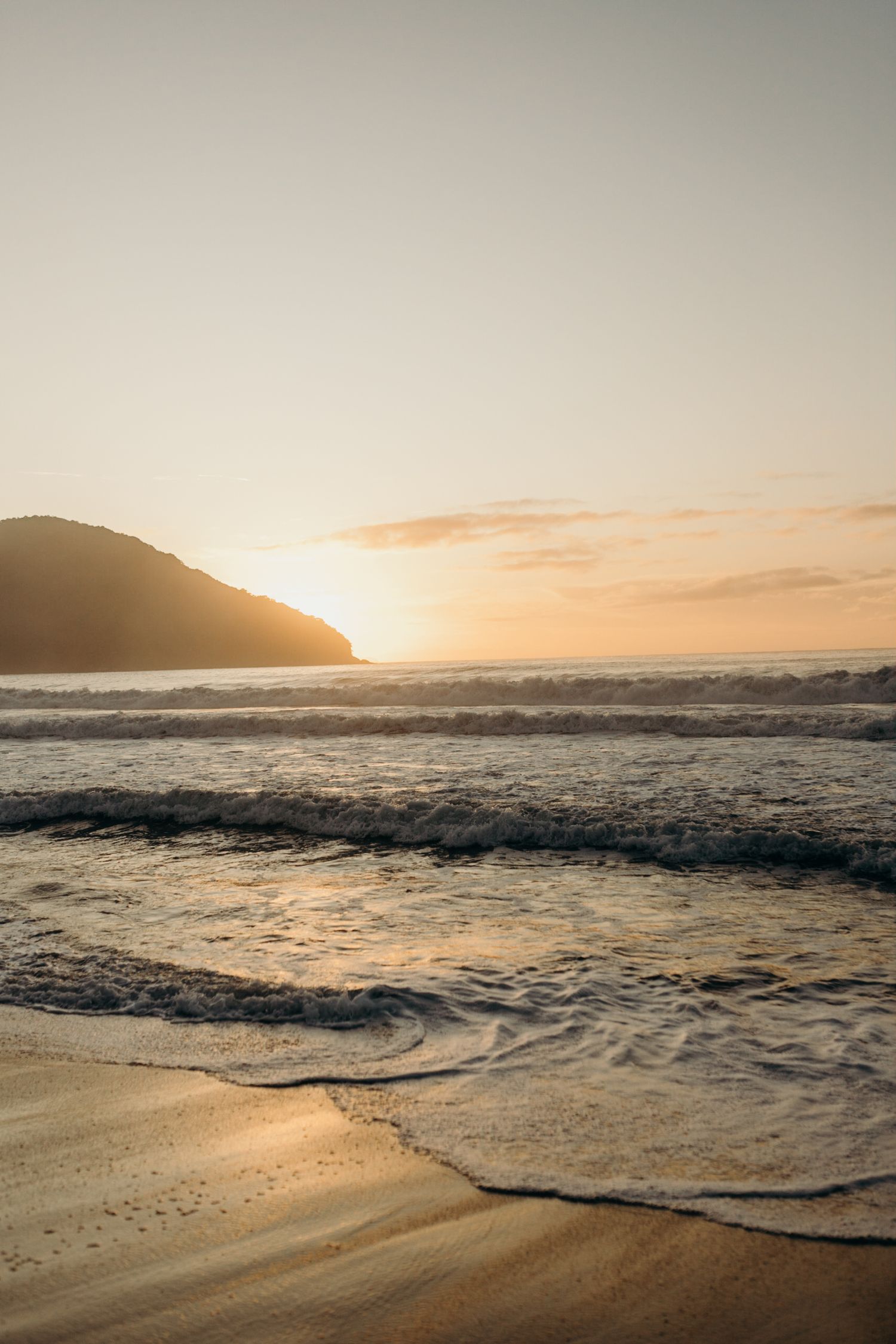 Sunset over mountain silhouette with waves crashing on sandy beach during golden hour.