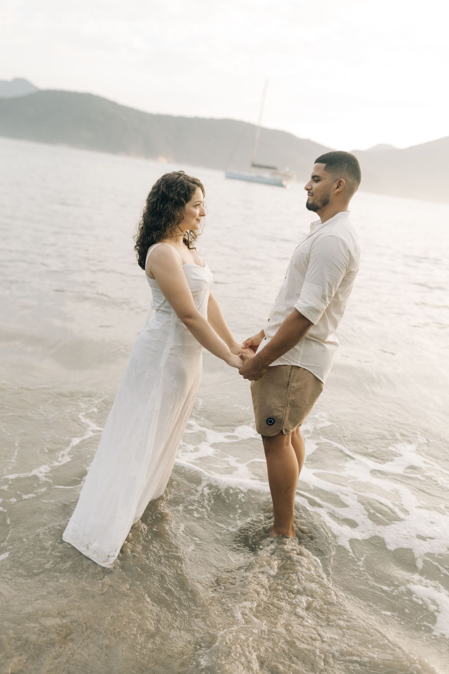 A couple holds hands while standing in ocean waves with mountains in the background.
