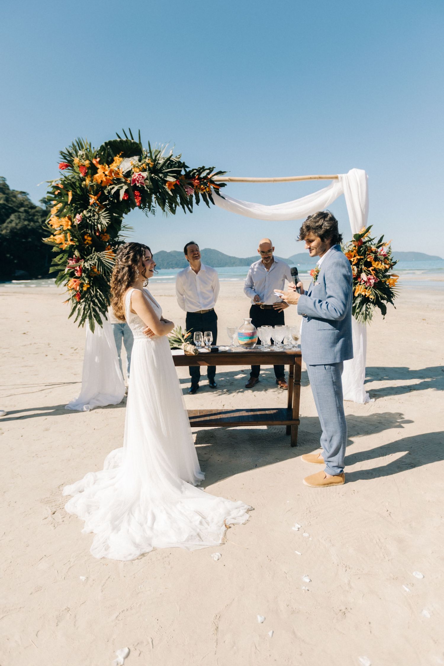 Beach wedding ceremony setup with floral arch and benches against ocean backdrop.