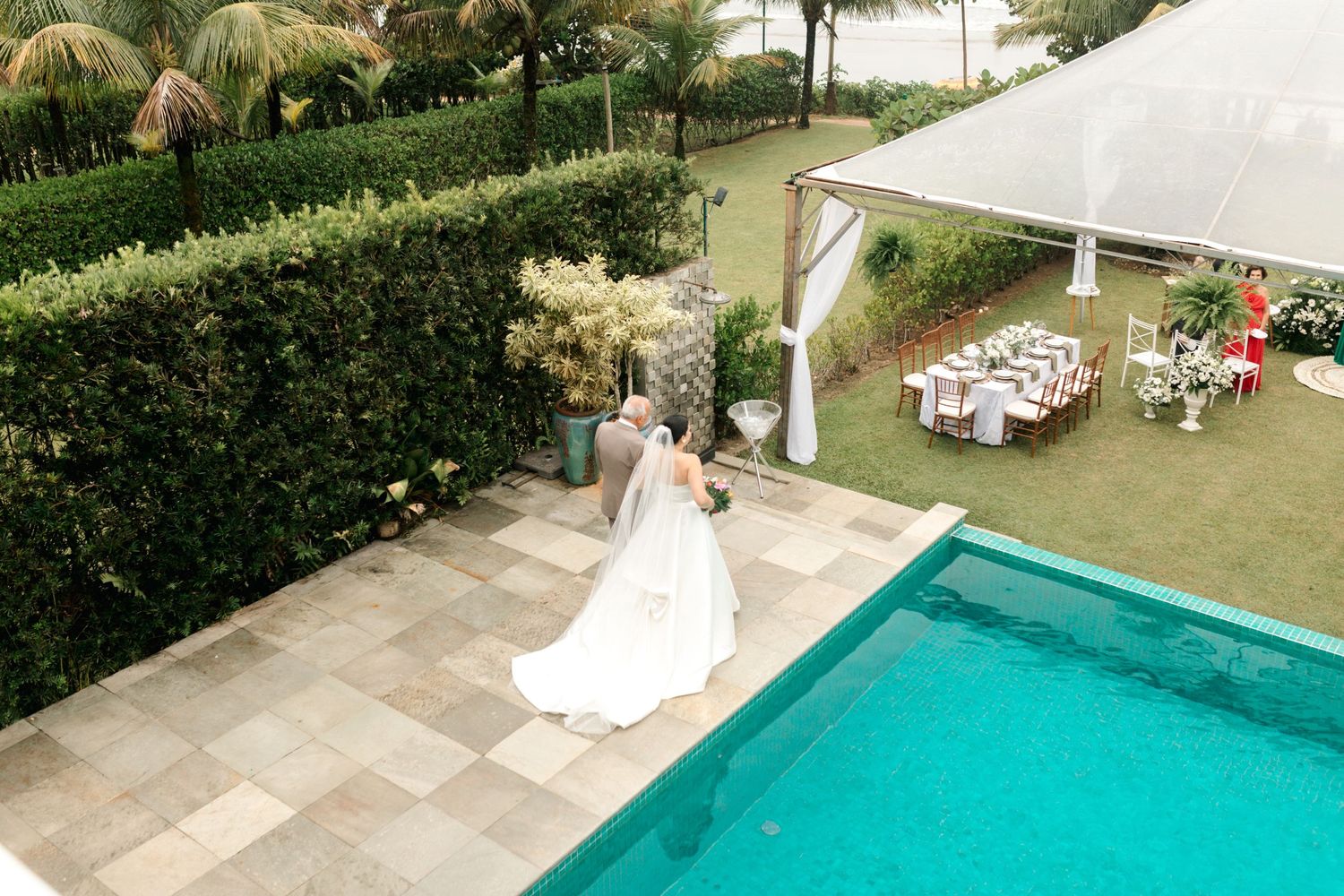 Aerial view of wedding ceremony setup with pool, white tent, and tables in a lush garden setting.