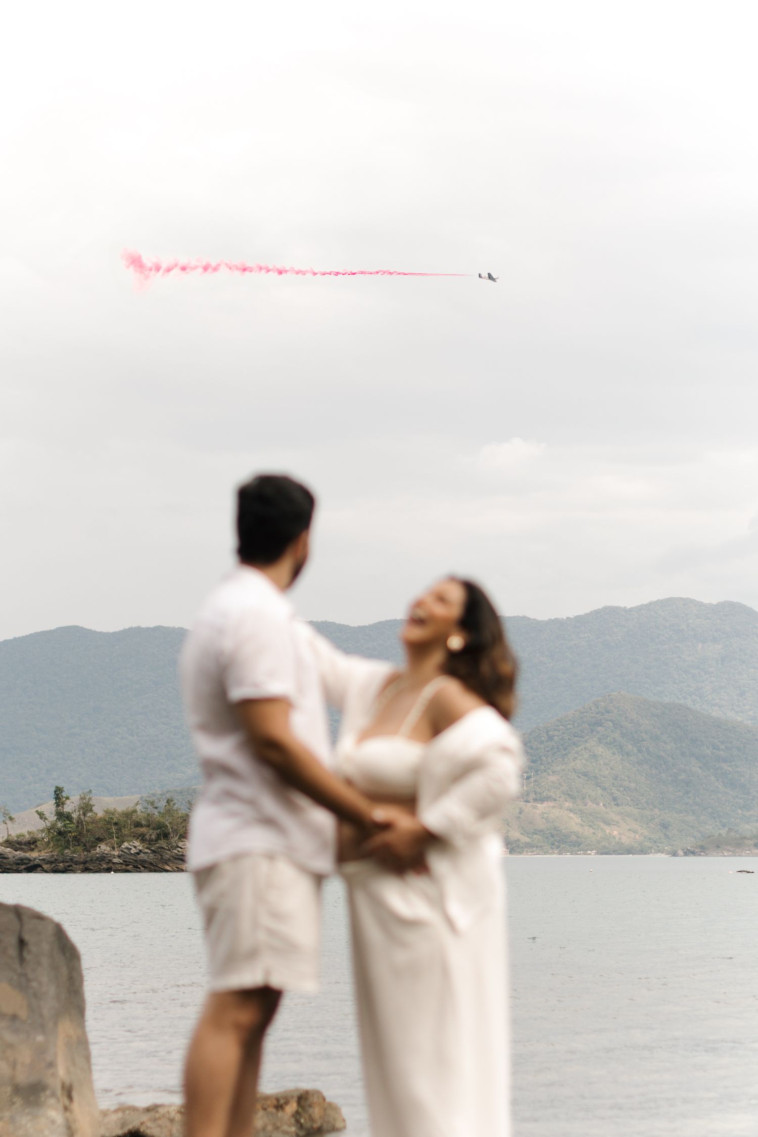 A couple in white attire embraces romantically by the ocean with mountains in the background.