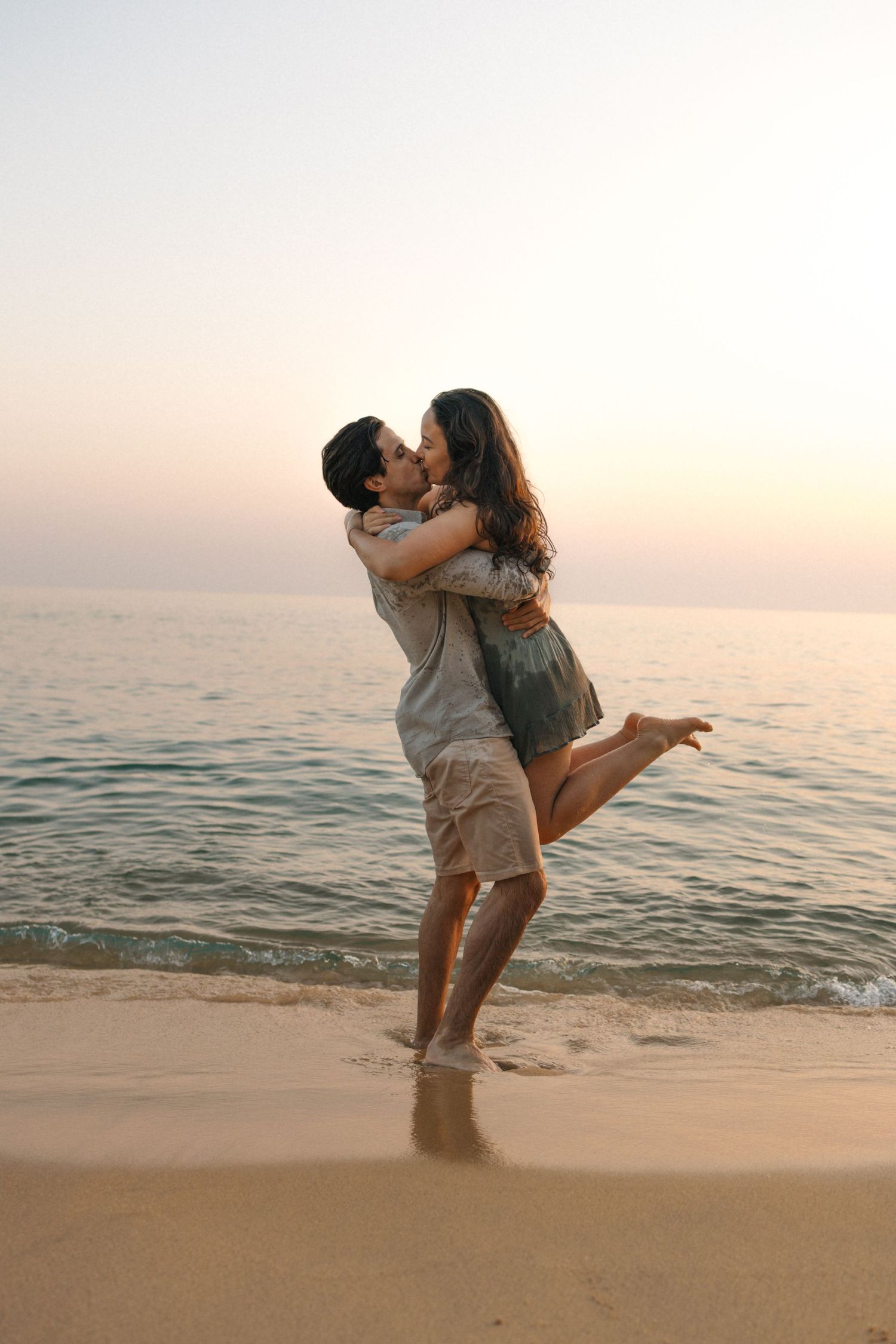 A romantic couple embraces and dances together on a sandy beach at sunset with ocean waves in the background.