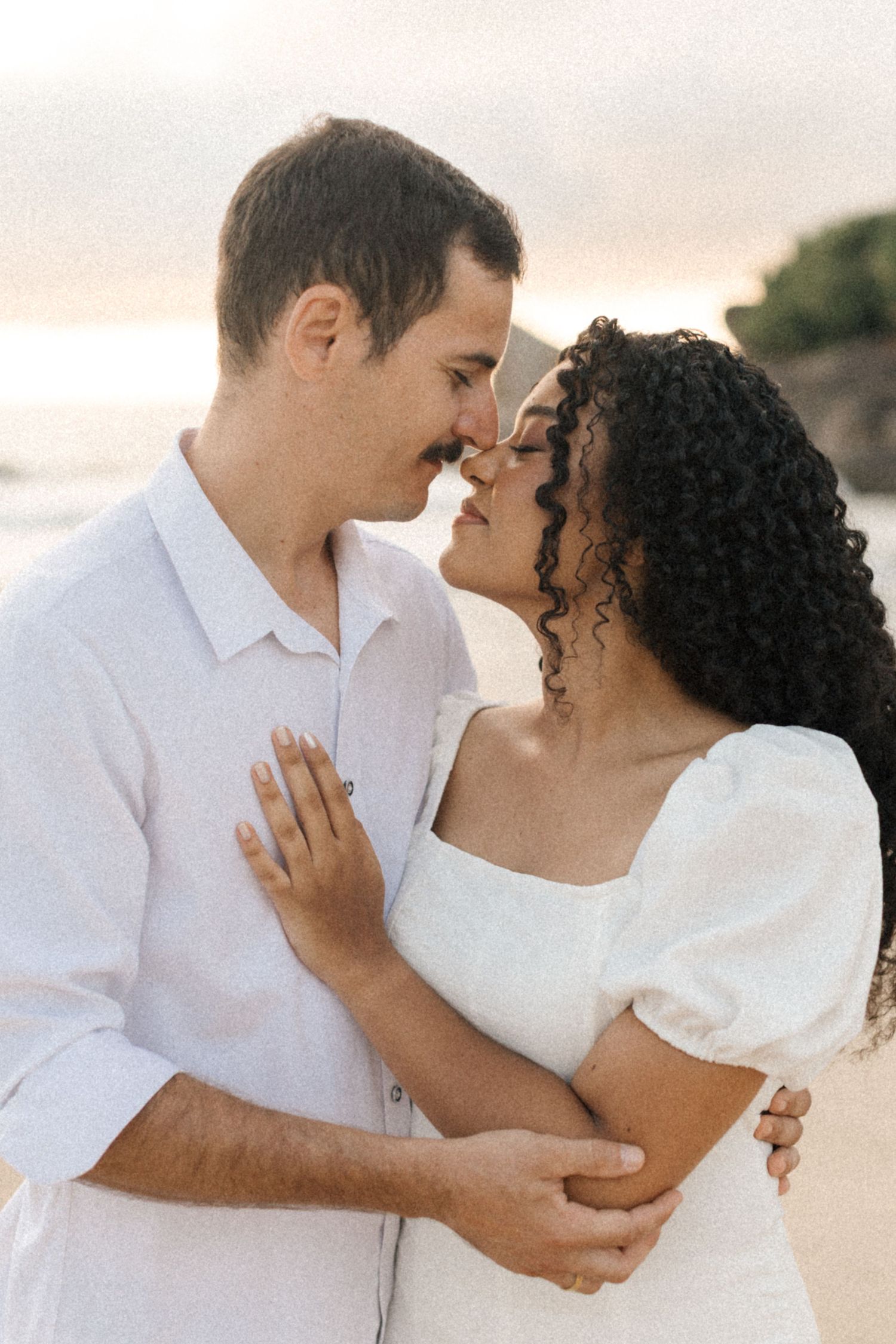 A loving couple in white outfits share intimate embraces during a romantic beach photo session at sunset.