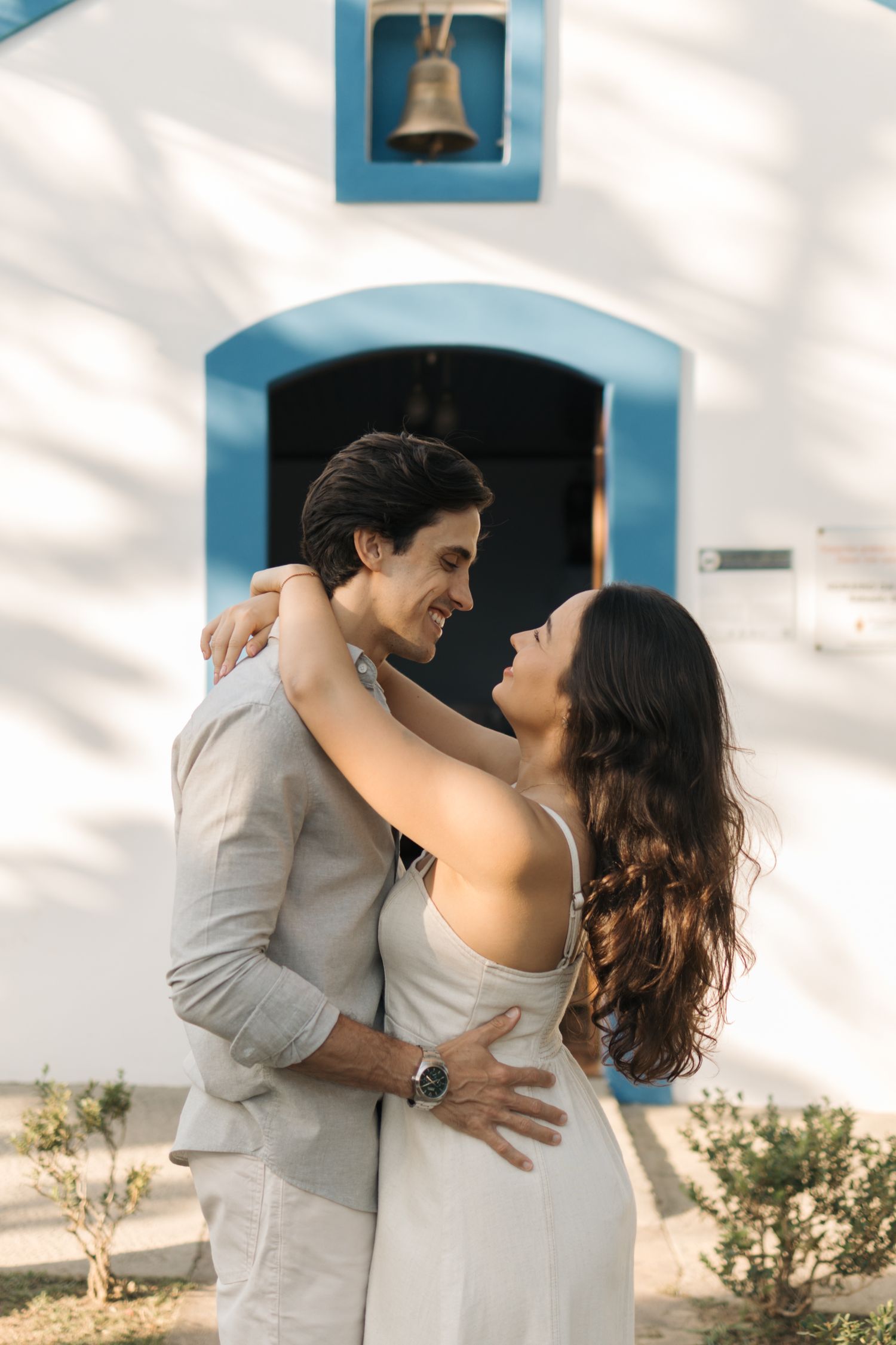 Romantic couple embraces and laughs together against white church wall with blue accents during sunset photoshoot.