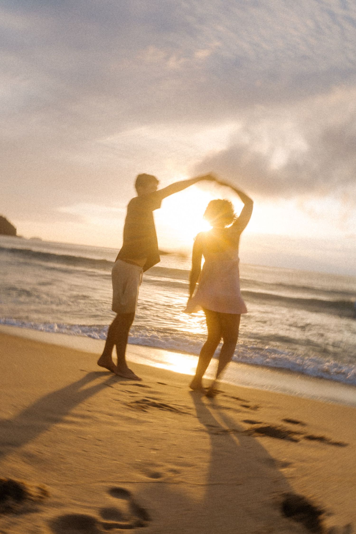 Duas silhuetas dançam juntas em uma praia ao pôr do sol enquanto a luz dourada flui através de suas formas dançantes.