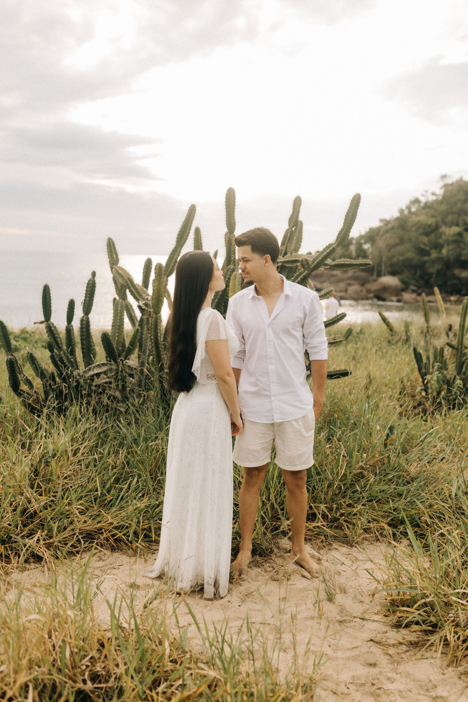 A couple in white clothing stands among cacti in a coastal landscape setting.