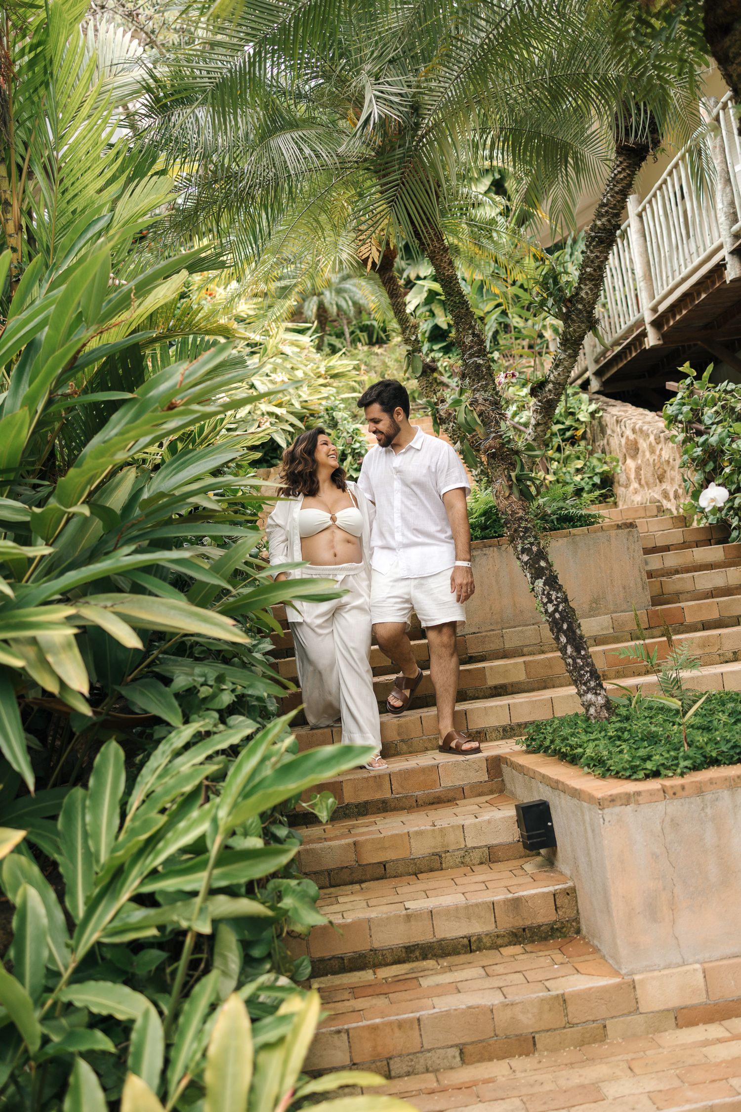 Two people in white outfits descending outdoor brick stairs surrounded by lush tropical plants and palm fronds.